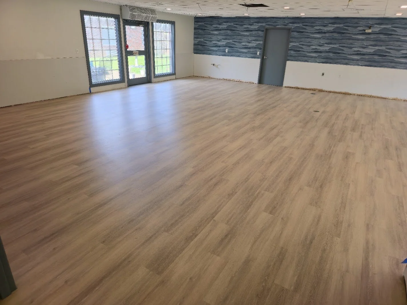Empty room with light wooden flooring, large windows and door at the front, blue textured wall on the right, and plain white walls.