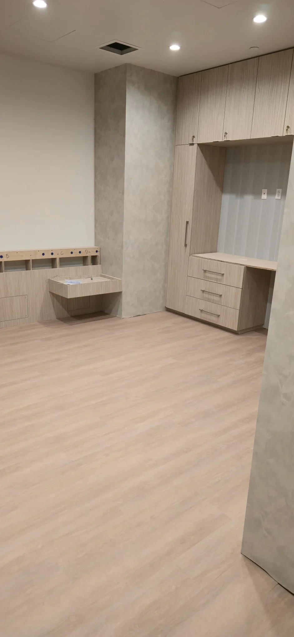 An empty room with light-colored wood flooring and built-in cabinetry, including a tall cabinet and drawer unit, with a small floating shelf and a wall panel with labeled holes.