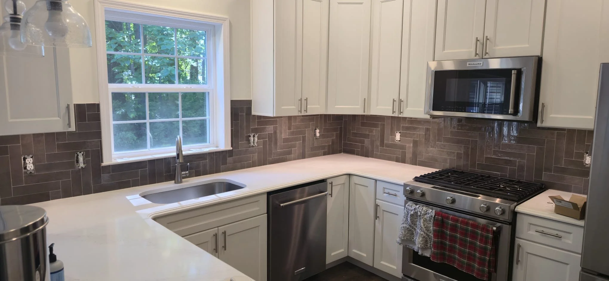 An unfinished kitchen with white cabinets, a window with green trees outside, a grey tiled backsplash, a sink, and stainless steel appliances.