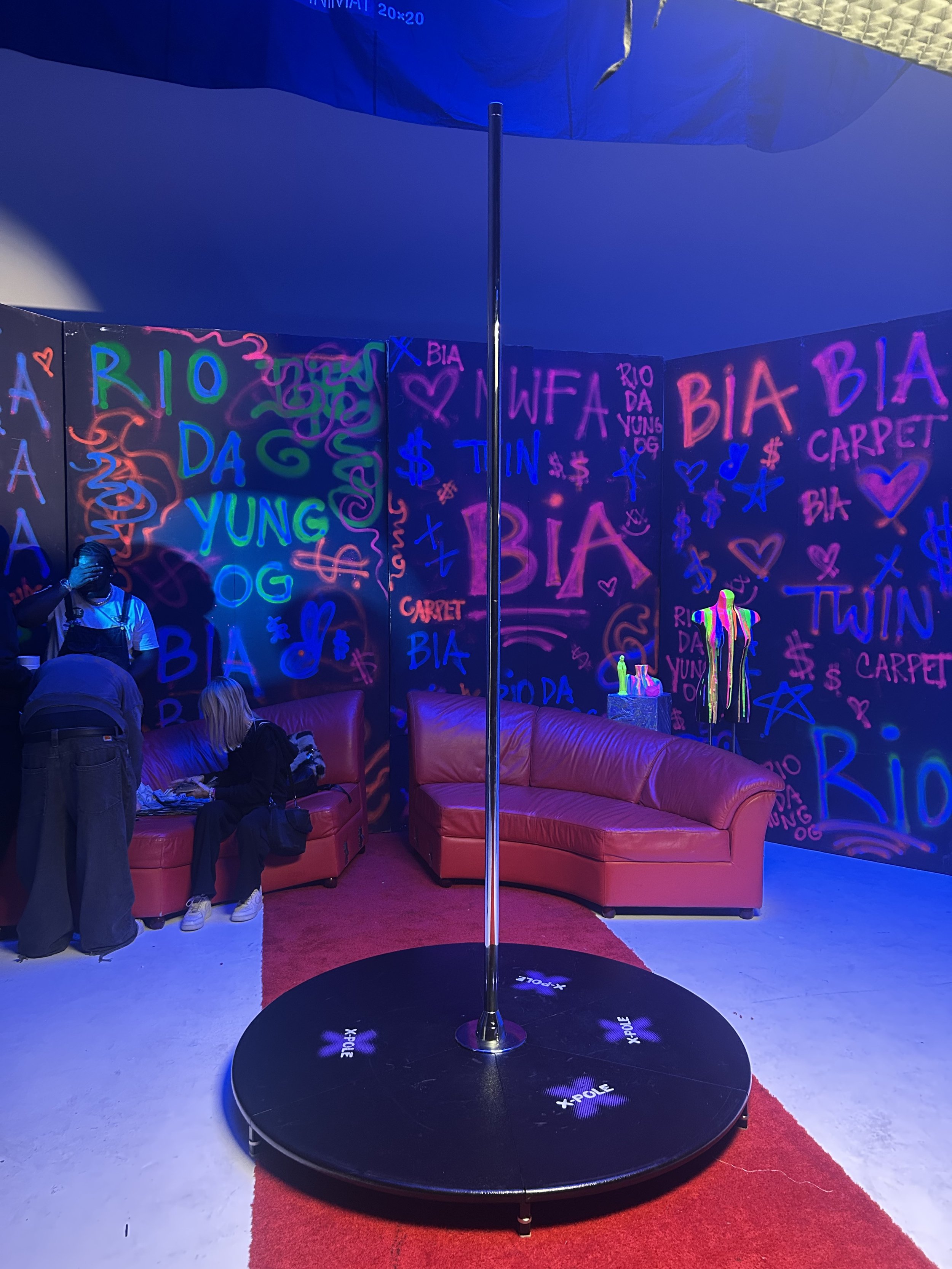 An indoor event space with red couches, a small dance floor with a pole in the center, and walls decorated with neon-style words and symbols in pink, blue, and green. The background features graffiti-style text that reads 'RIO DA YUNG OG', 'BIA', and other words and symbols.