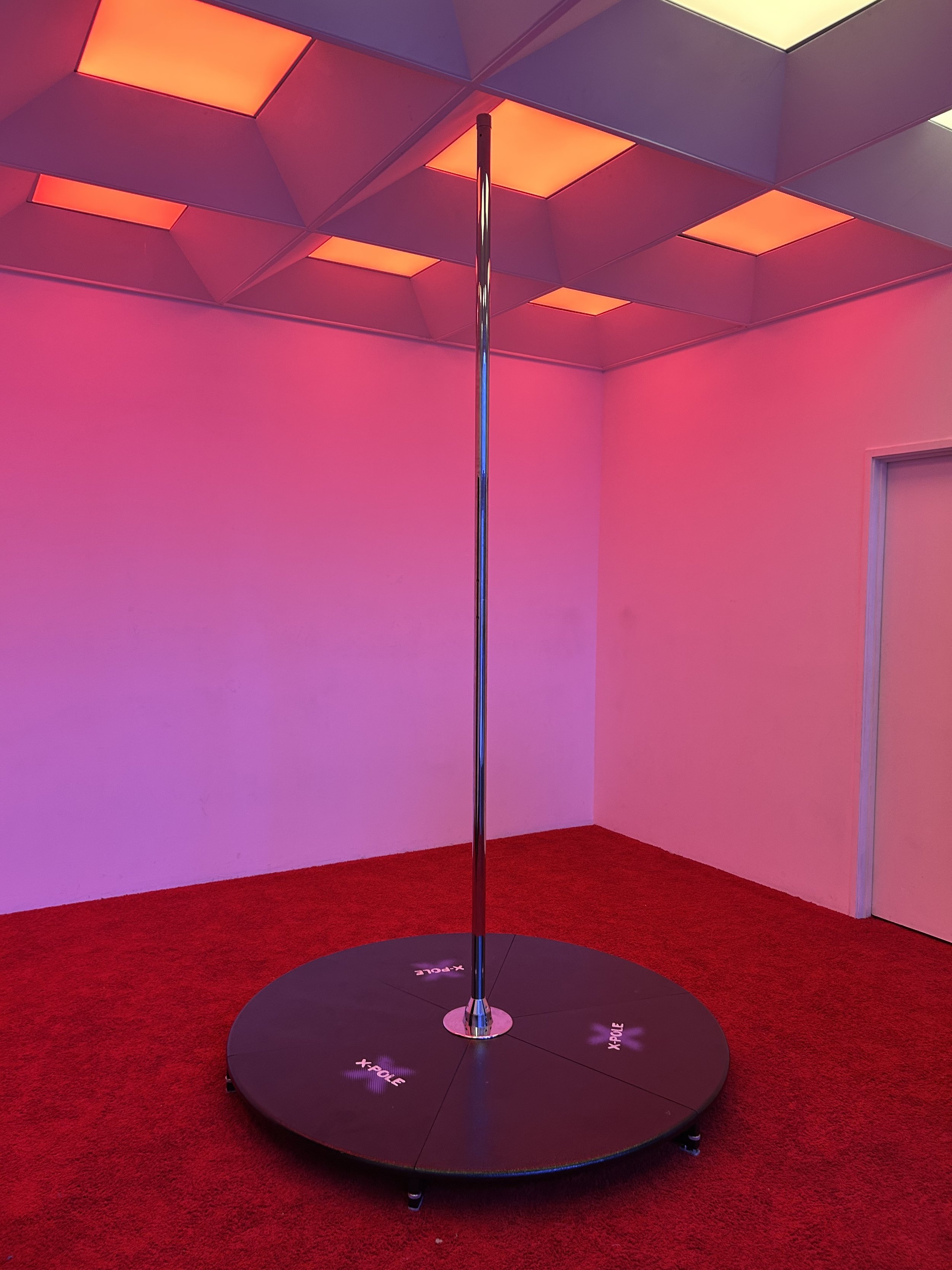 An indoor dance studio with a black circular dance platform and a chrome stage pole in the center, illuminated by colorful overhead lights, with red carpeted floor and pink walls.
