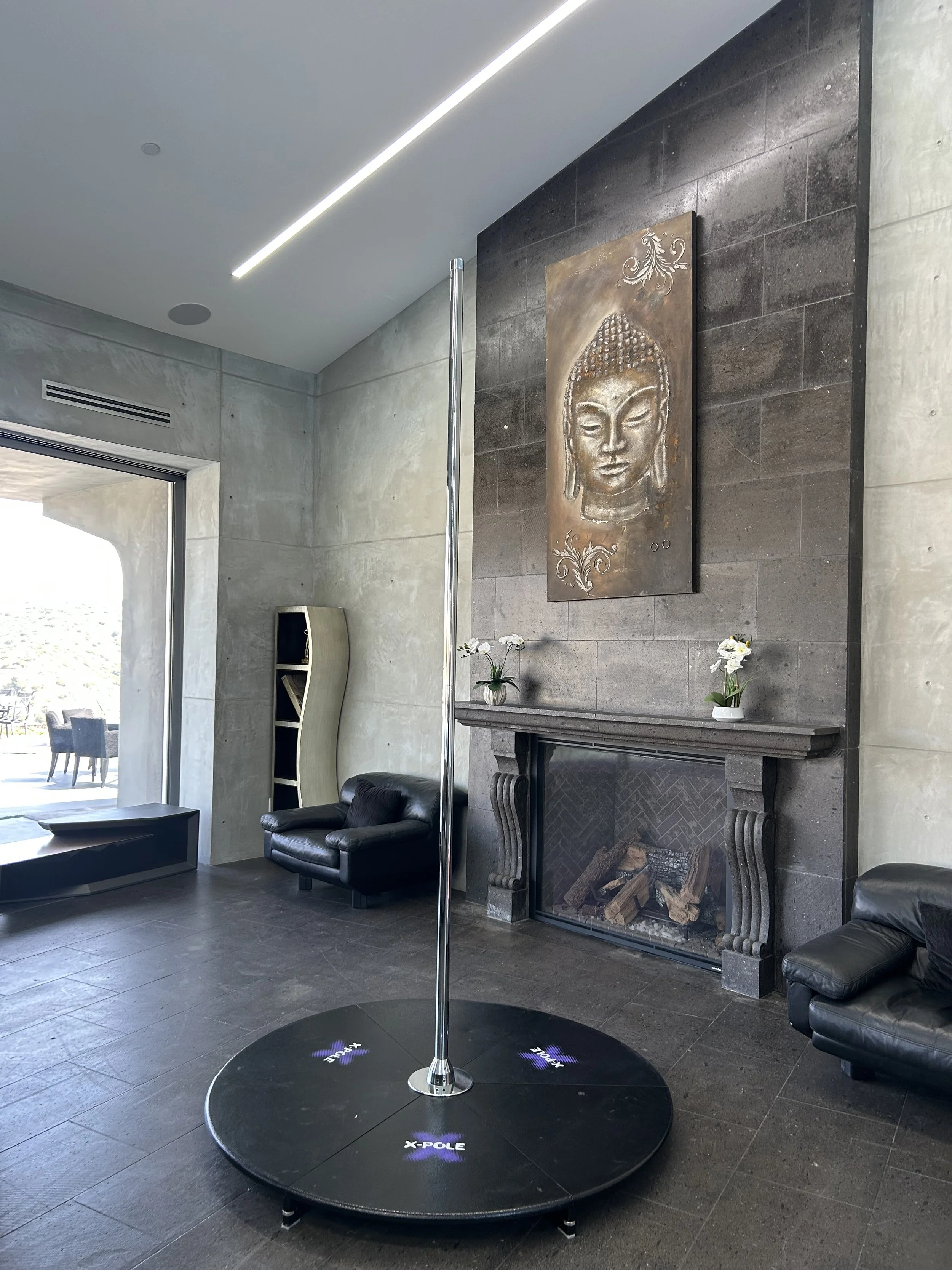 Indoor space with modern decor, featuring a black leather armchair, a large art piece of Buddha's face on the wall, potted orchids on a mantle, a black turntable with a chrome stage pole, and a window with an outdoor seating area.