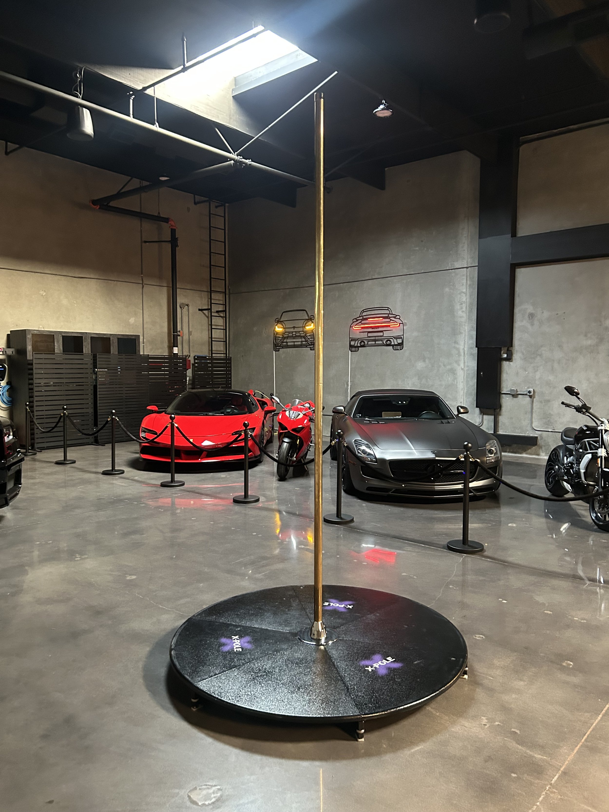 A small indoor car showroom with several luxury sports cars, including red and silver models, and a brass stage pole at the center on a black circular base with orca whale logos.