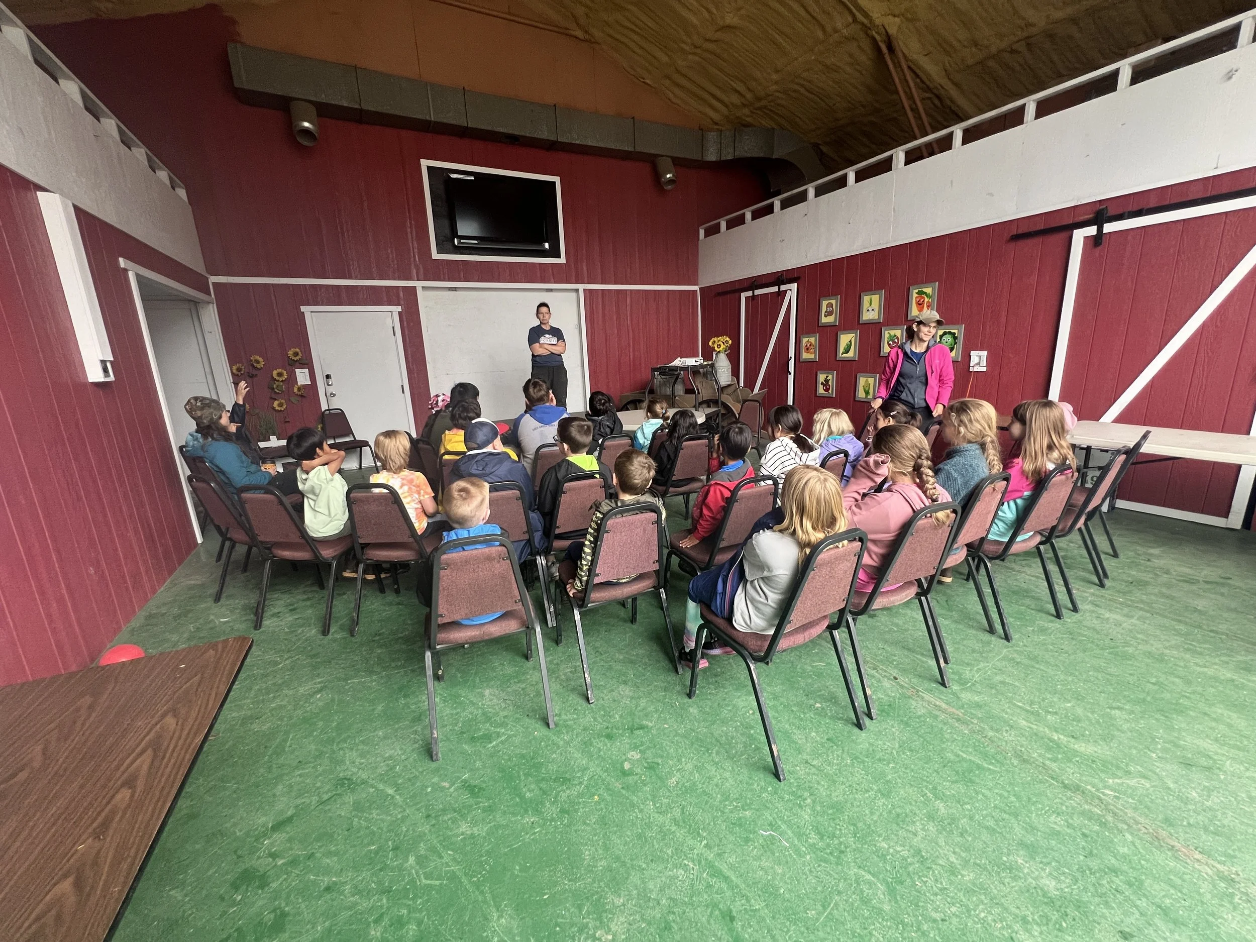 Children seated in chairs listening to a woman speaker in a room with red and white walls decorated with framed pictures, on green floor.