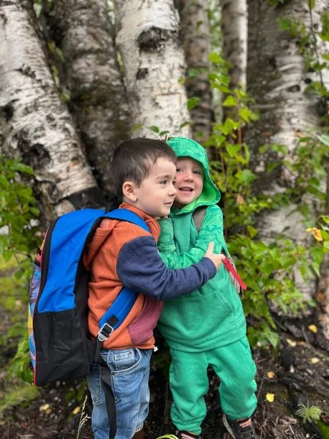 Two young children, one boy and one girl, hugging each other outside in front of a forest with birch trees, surrounded by green leaves.