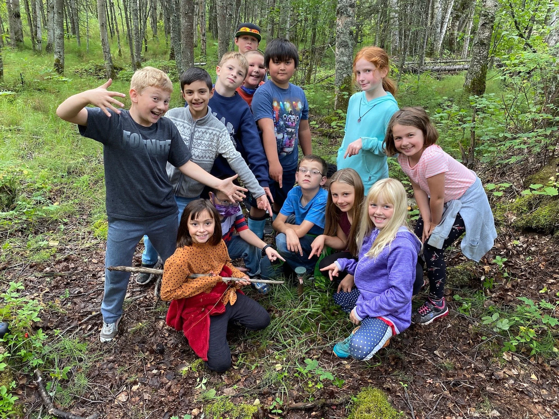 A group of children posing in a forest, some kneeling and some standing, surrounded by trees and green foliage.