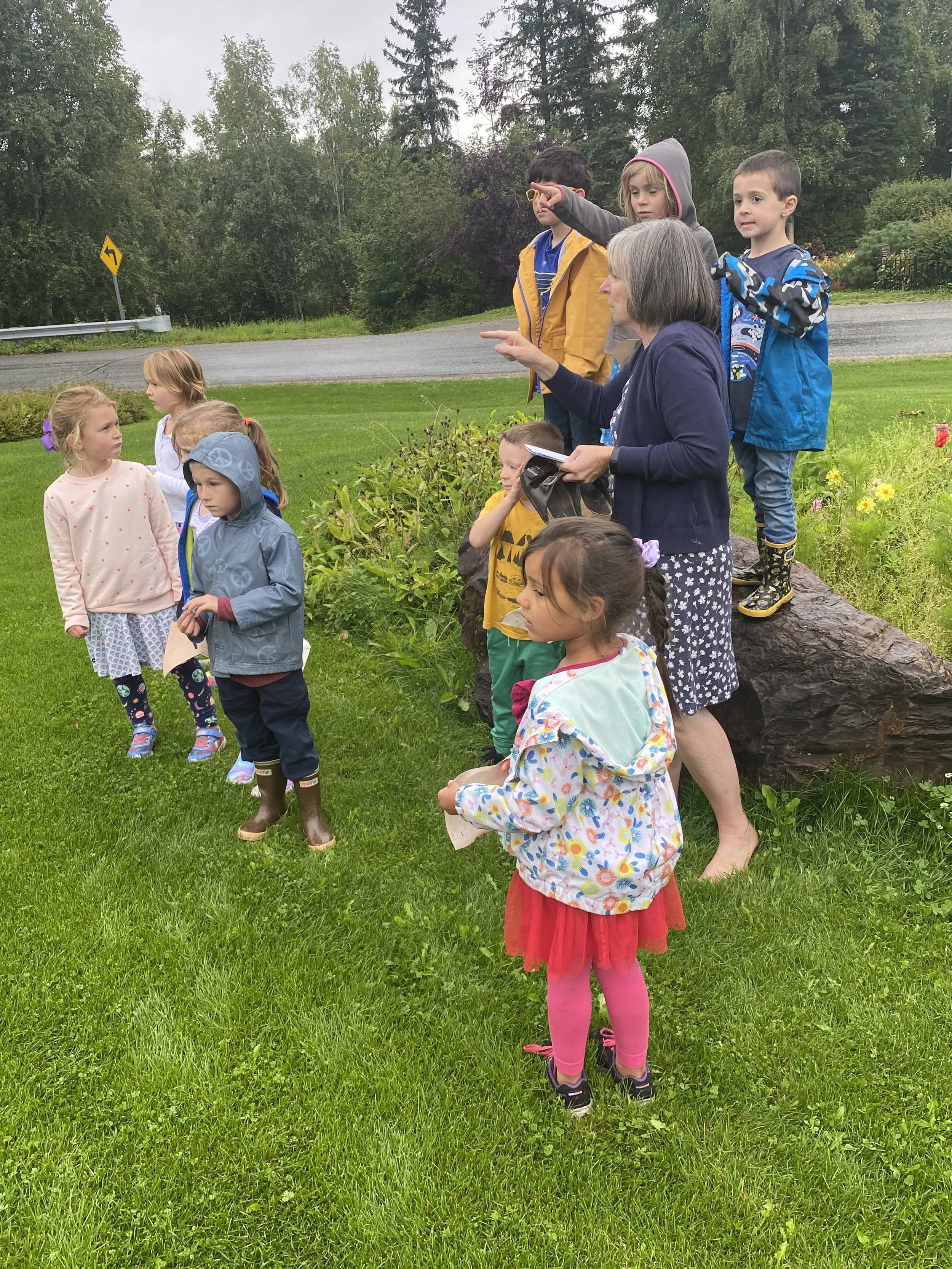 A group of children and a woman standing outdoors on a grassy area near a road, with trees in the background. The woman is pointing at something, and the children are looking in the same direction, wearing rain jackets and casual clothing.