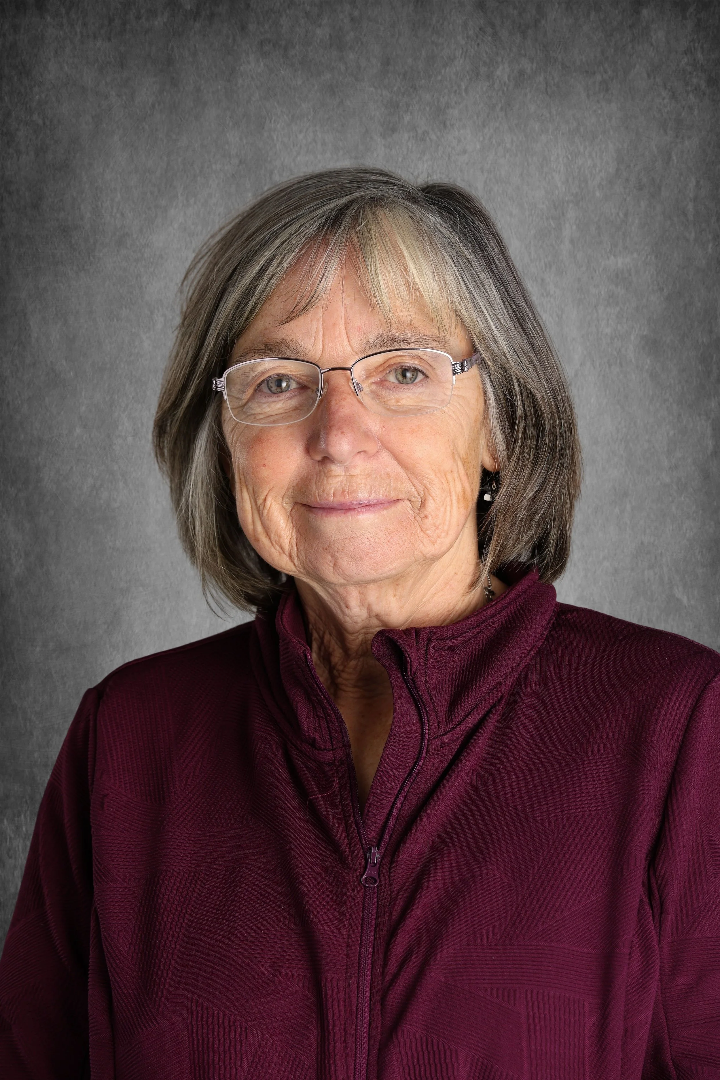 An elderly woman with shoulder-length gray hair, wearing glasses and a maroon zip-up jacket, against a gray textured background.