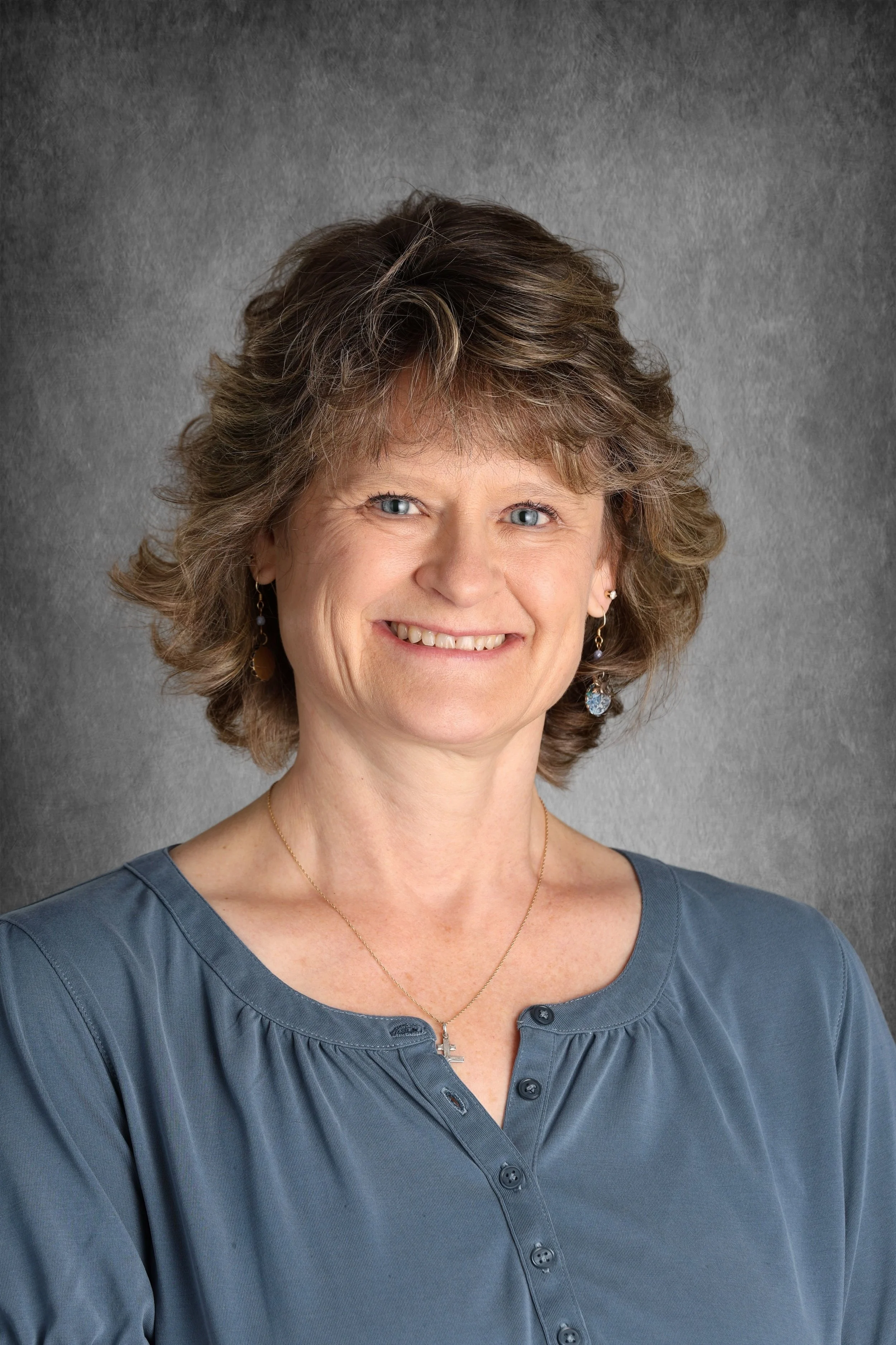 A smiling woman with short brown hair and blue eyes, wearing a blue top, earrings, a gold necklace with a cross pendant, and a matching chain, standing in front of a gray background.