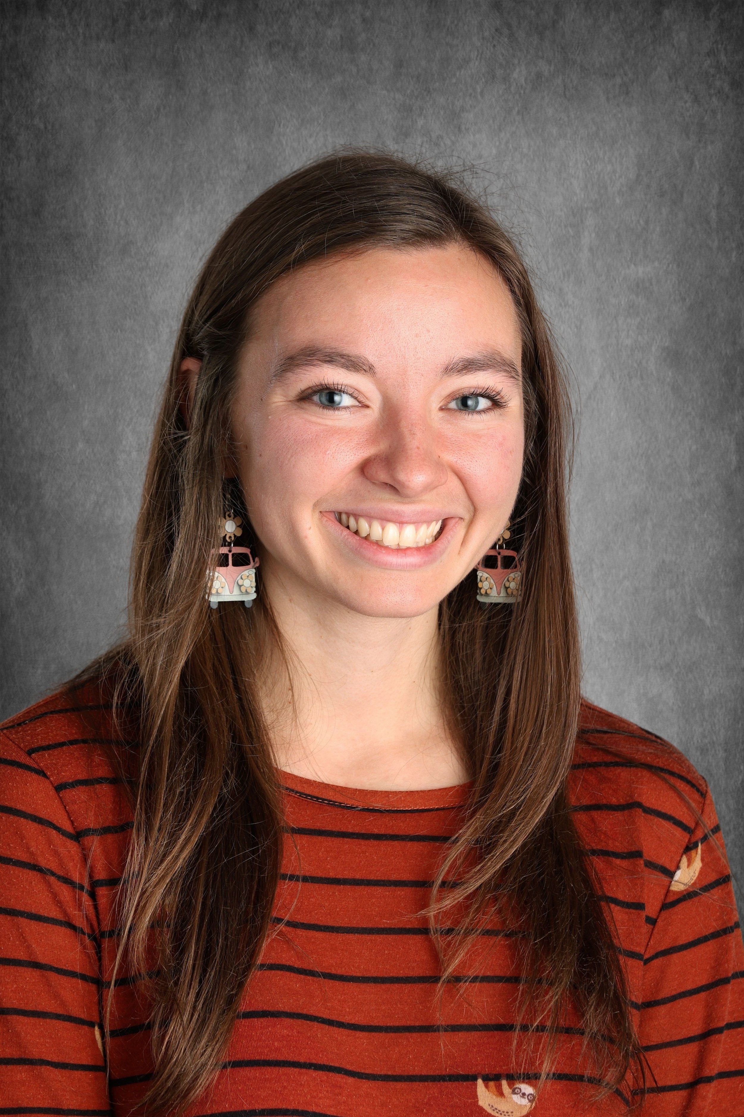 A young woman with long brown hair and blue eyes smiling, wearing earrings with a house design, a striped orange and black shirt, in front of a gray textured background.