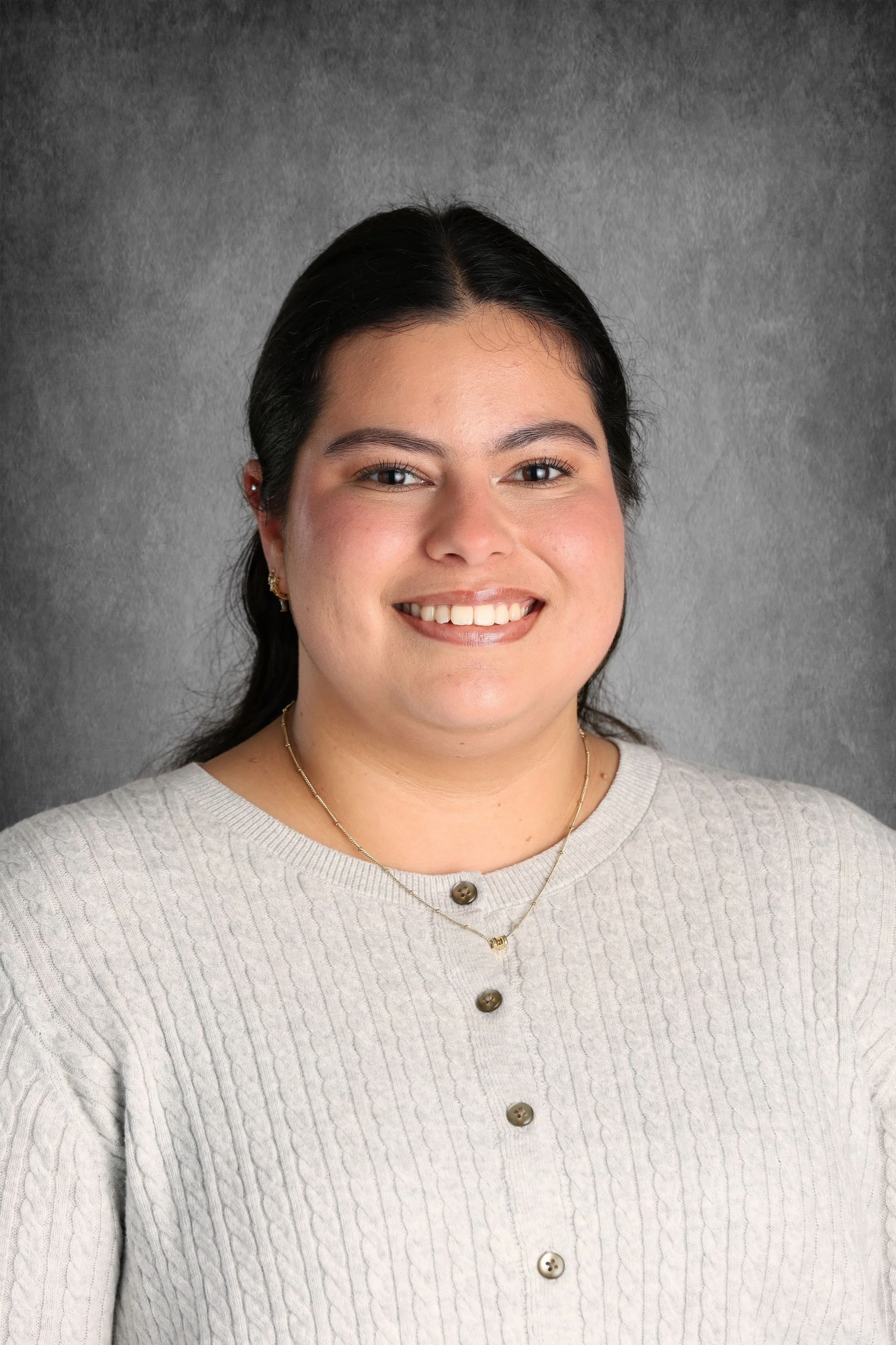 A woman with shoulder-length dark hair and light skin, smiling in front of a gray textured background, wearing a light-colored, cable-knit sweater with large buttons and gold jewelry.
