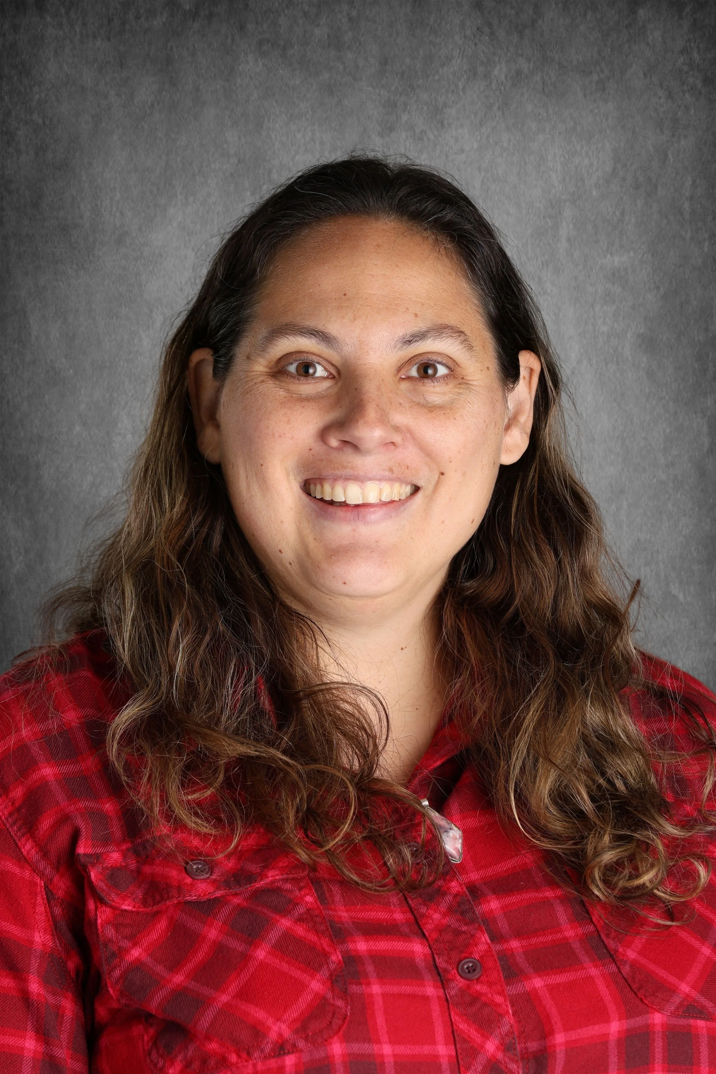 Portrait of a woman with long, curly brown hair smiling at the camera, wearing a red plaid shirt, against a gray background.