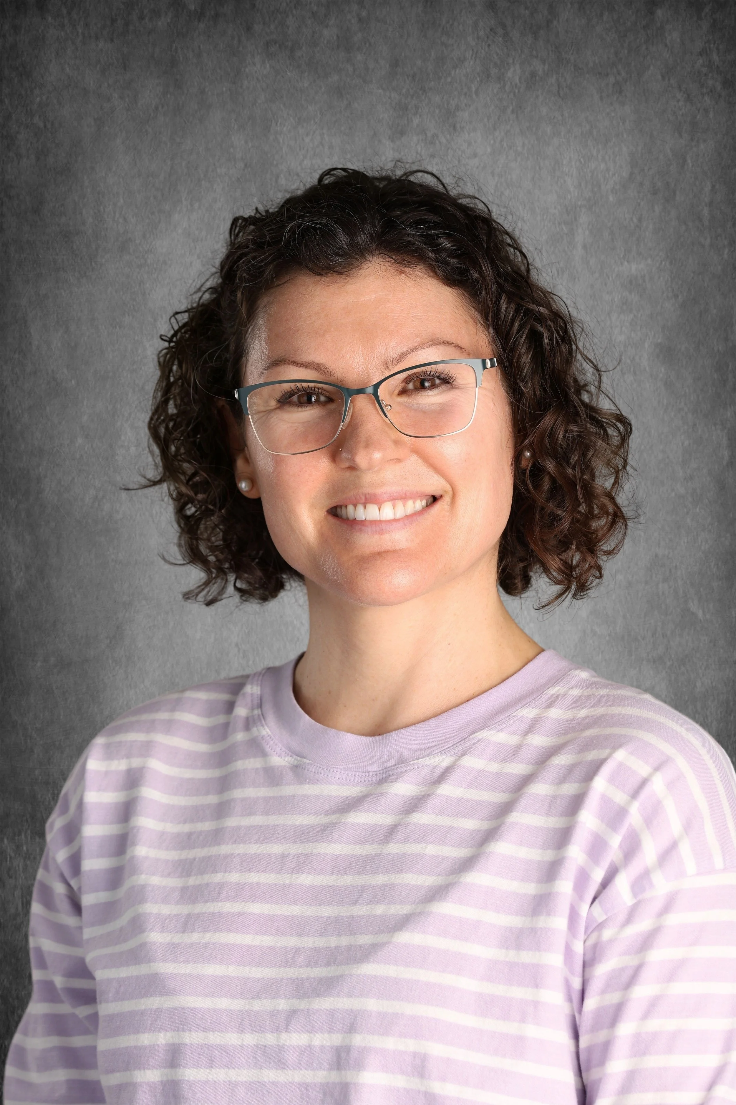 A woman with curly hair, glasses, and pearl earrings, smiling, wearing a light purple and white striped shirt, against a gray textured background.