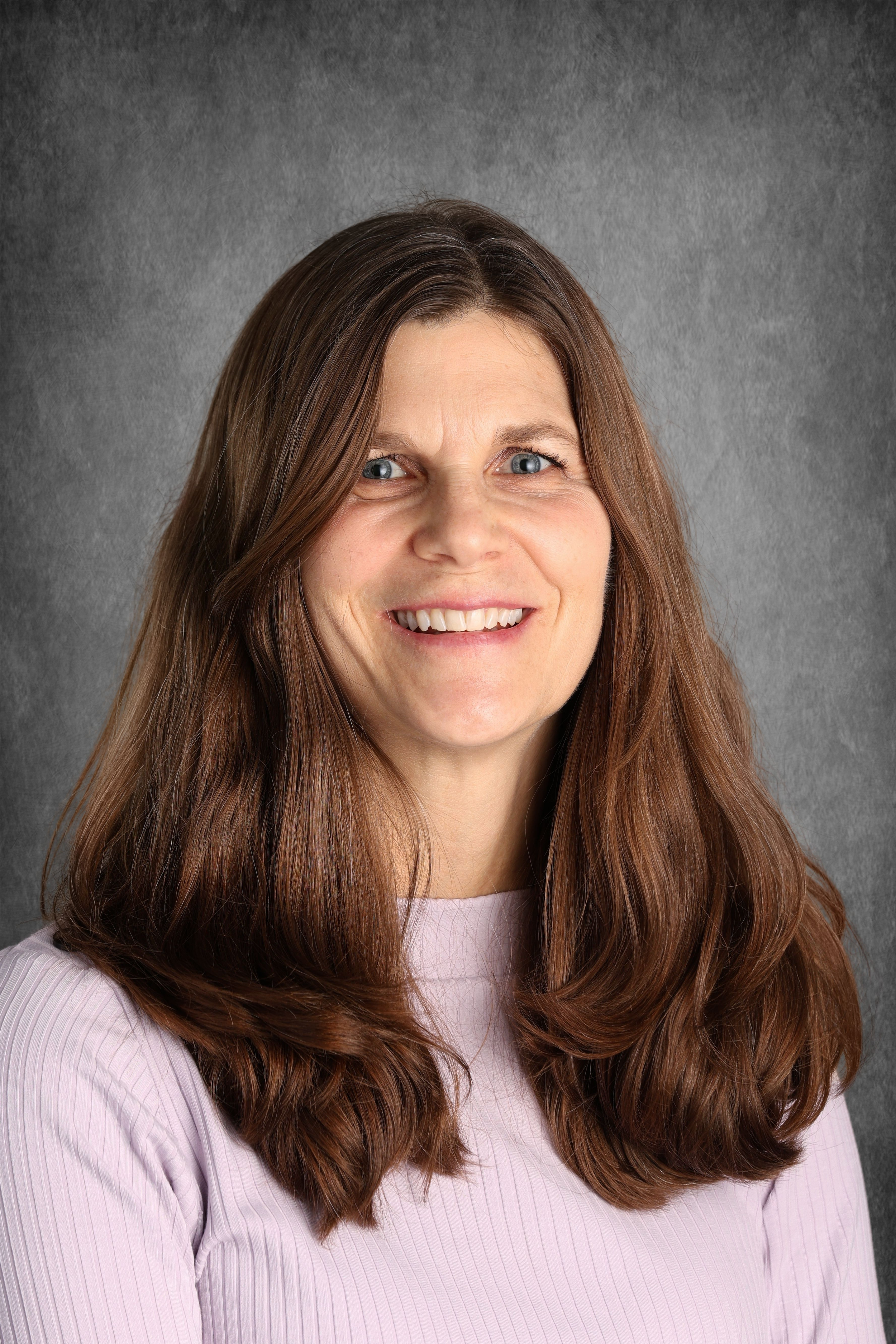 A woman with long, wavy brown hair and blue eyes is smiling at the camera, wearing a light pink, ribbed top against a grey textured background.