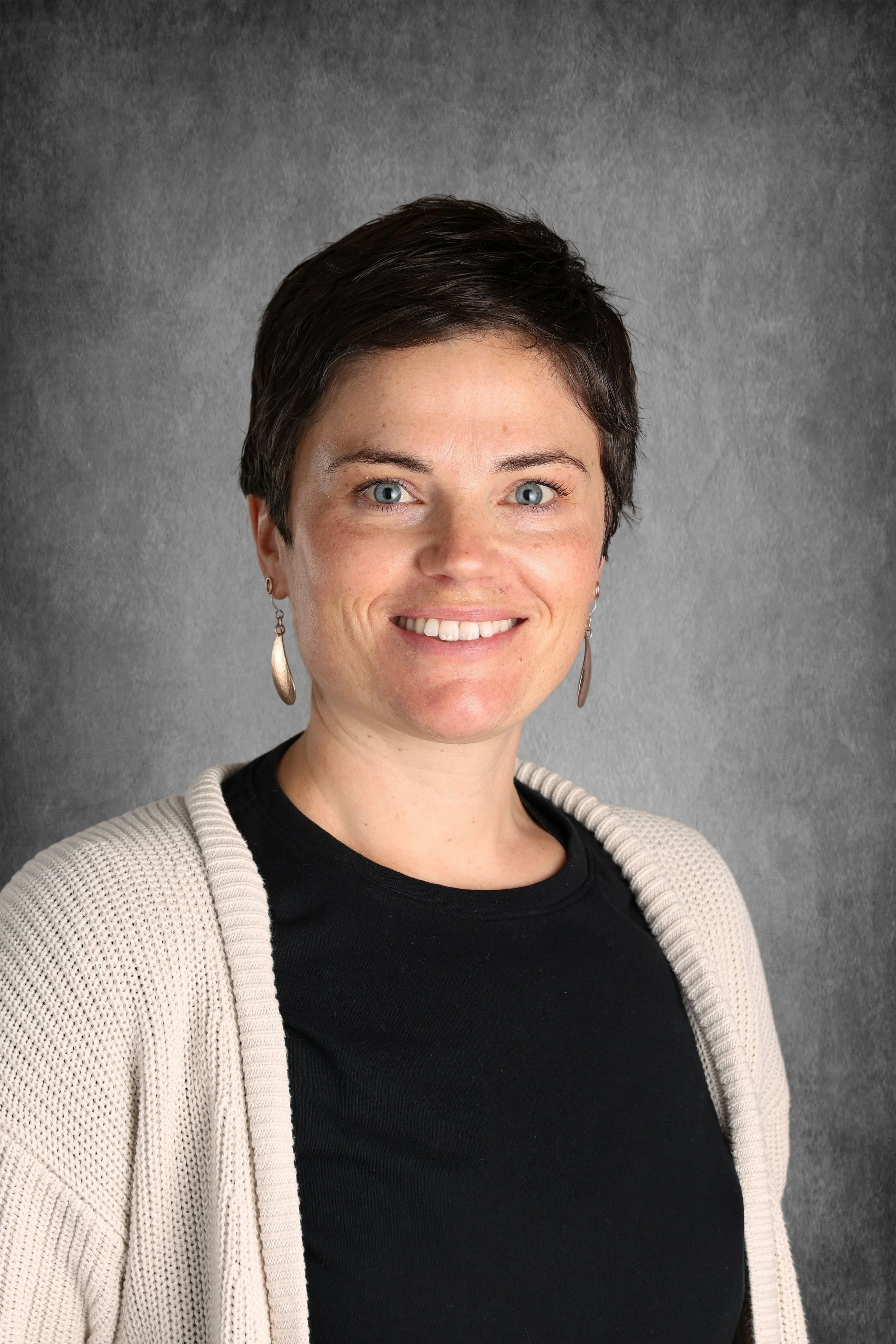 Headshot of a woman with short dark hair, blue eyes, wearing earrings, a black shirt, and a beige cardigan, standing in front of a gray background.