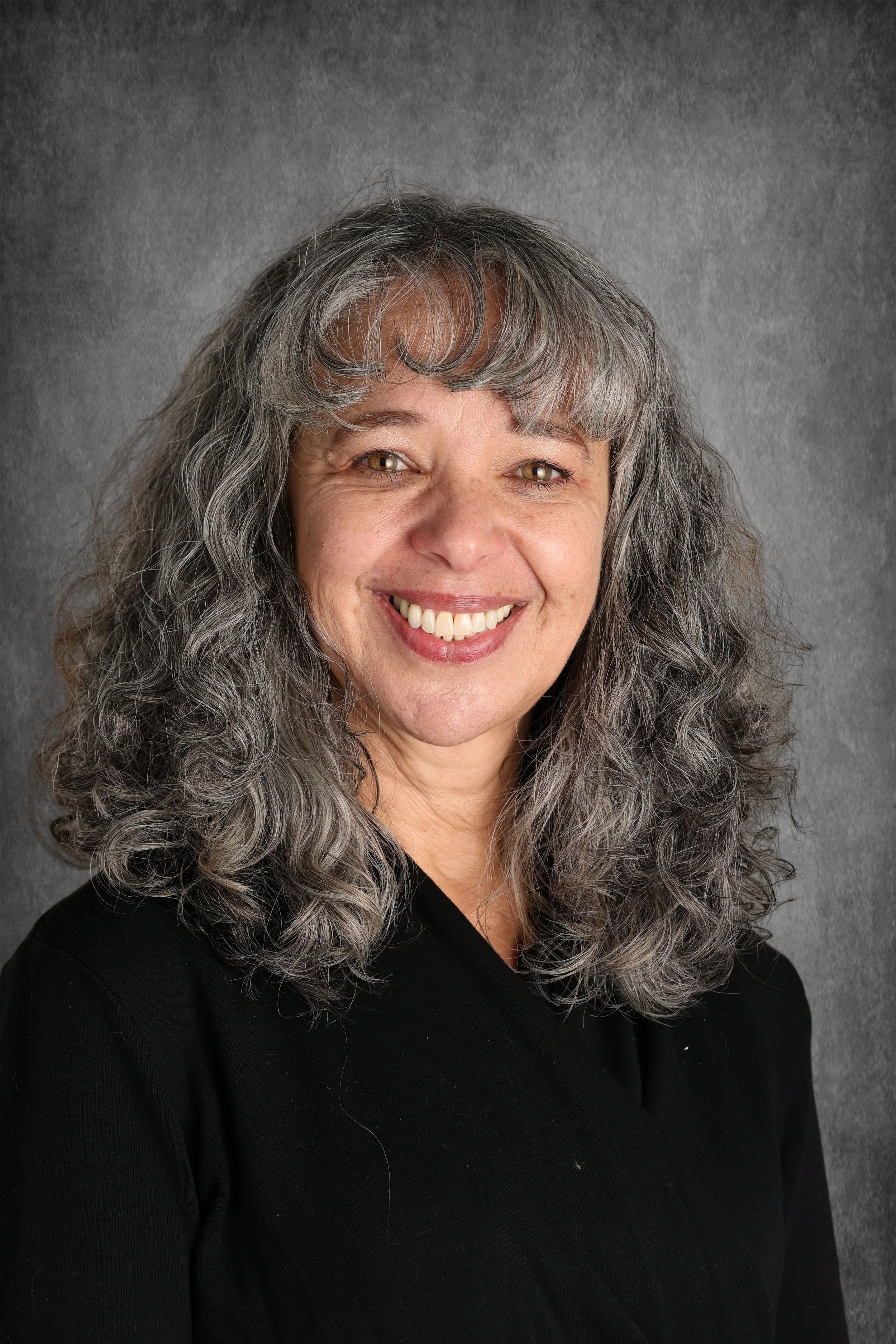 A woman with long curly gray hair, smiling, wearing a black top, against a gray textured background.
