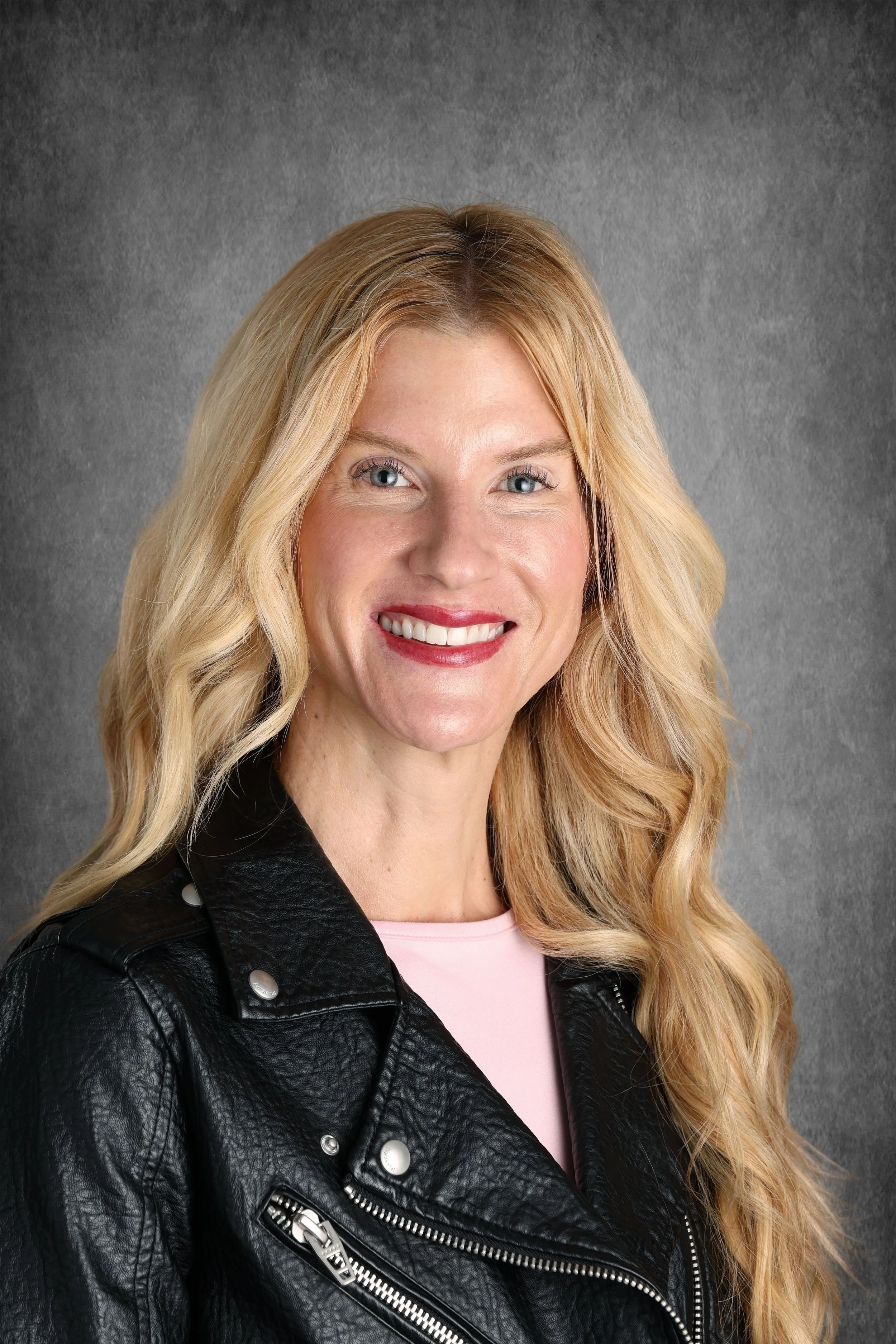 A woman with long blonde hair, blue eyes, wearing a black leather jacket and pink top, smiling at the camera against a gray background.