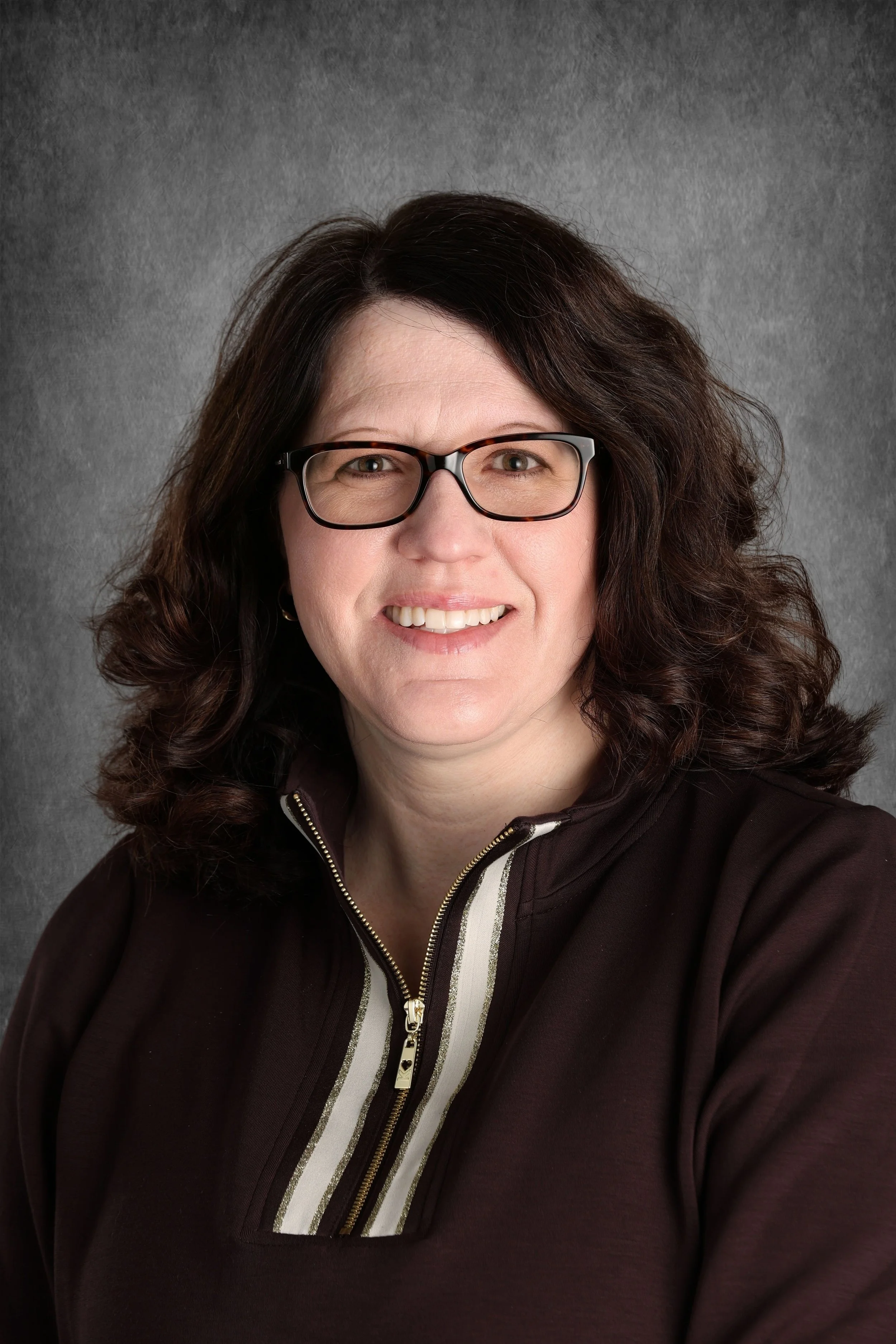 A woman with long, curly brown hair and glasses smiling at the camera, wearing a dark hoodie with a zipper and striped shirt underneath, against a gray textured background.