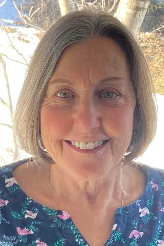 A smiling middle-aged woman with short gray hair, blue eyes, and a floral dress, standing outdoors in front of tree branches.