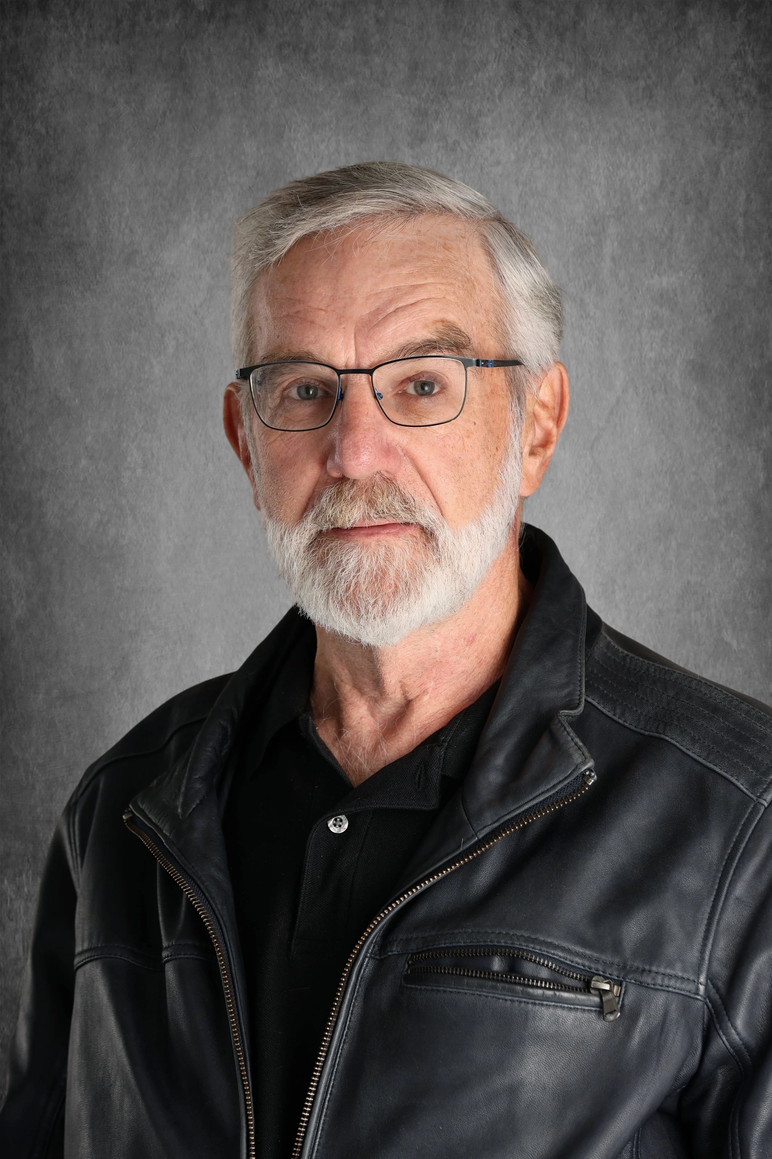 A close-up portrait of an older man with gray hair, a beard, and glasses, wearing a black leather jacket over a black shirt, against a gray textured background.