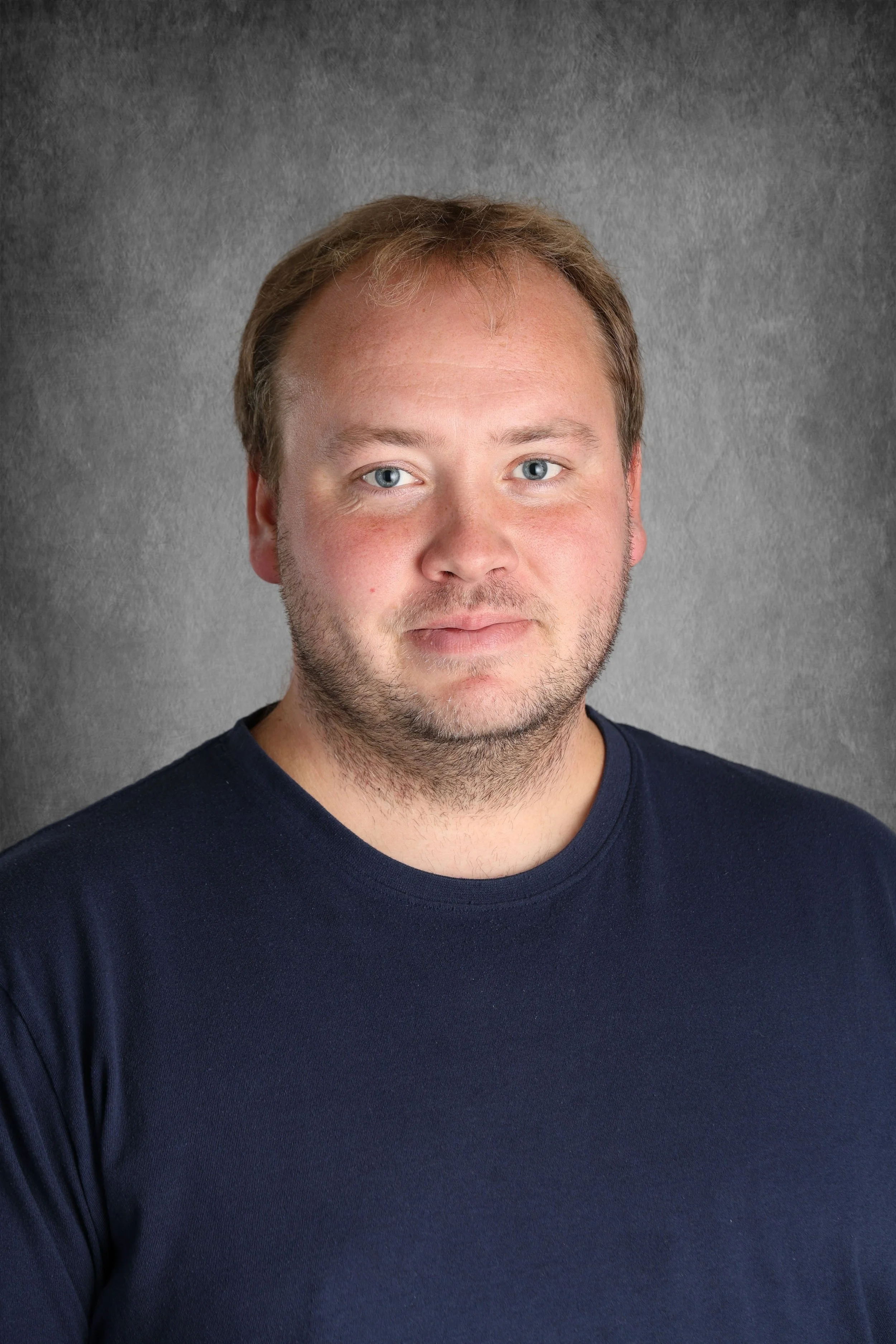 Close-up of a young man with light brown hair, blue eyes, a light beard, and mustache, wearing a navy blue T-shirt against a gray textured background.