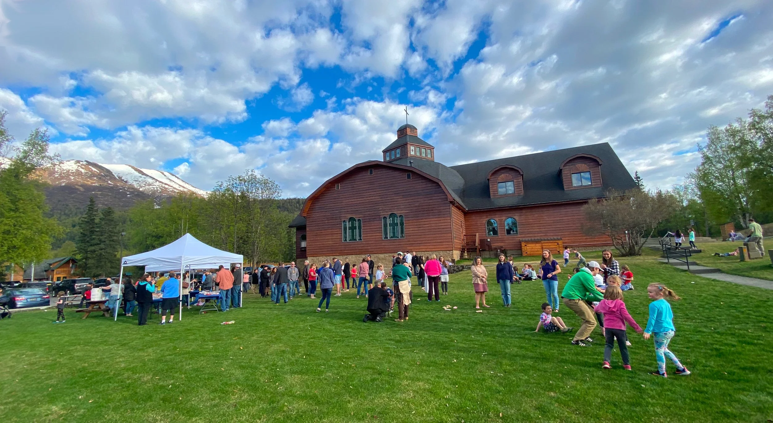 People gathered in front of a large wooden church on a grassy area, with a mountain and partly cloudy sky in the background.