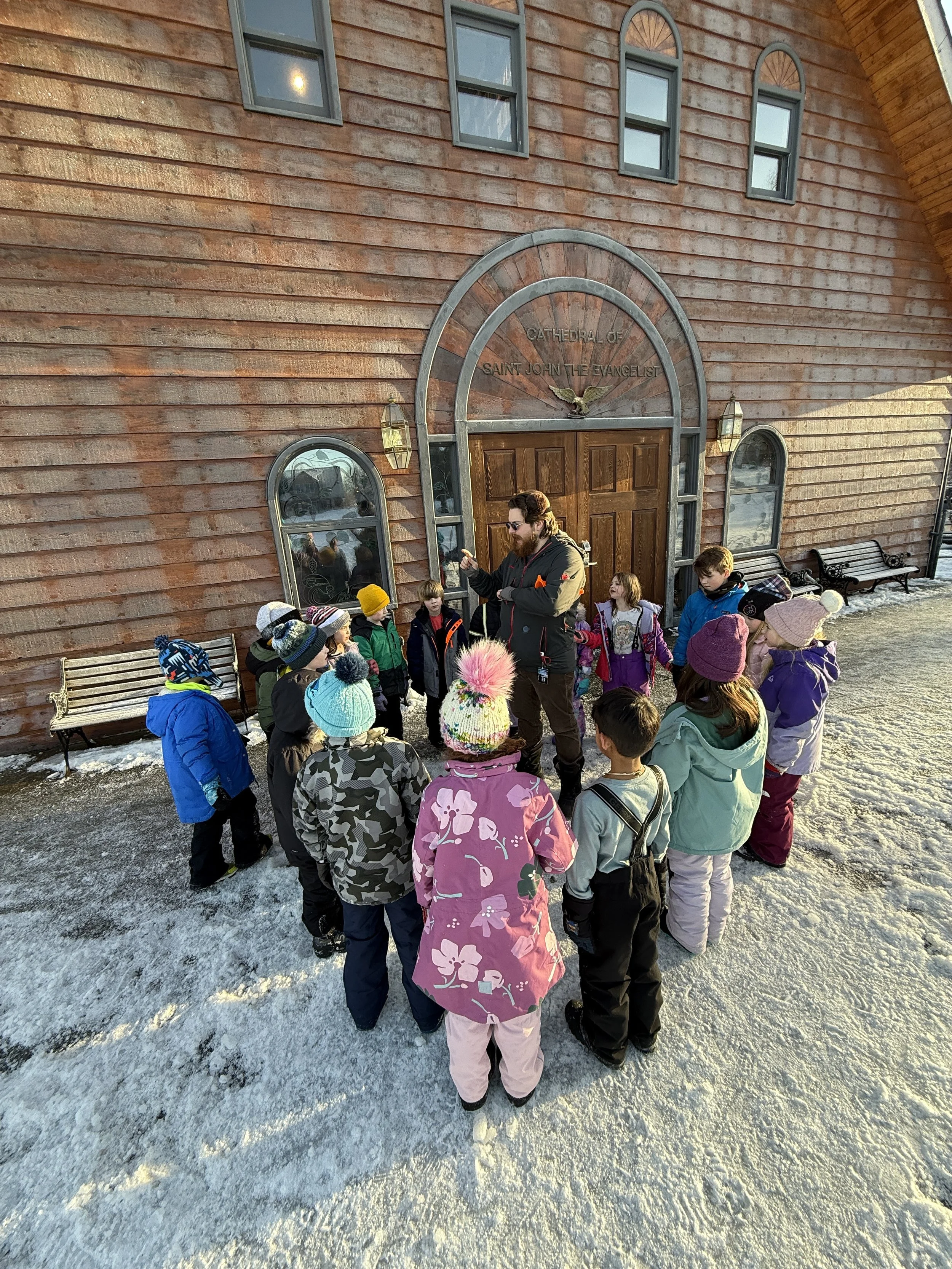 A group of children and a man standing outside a wooden church building with a sign that says 'Cathedral of Saint John the Evangelist'. The children are dressed in winter clothing, and the man appears to be giving a tour or speech to the children.