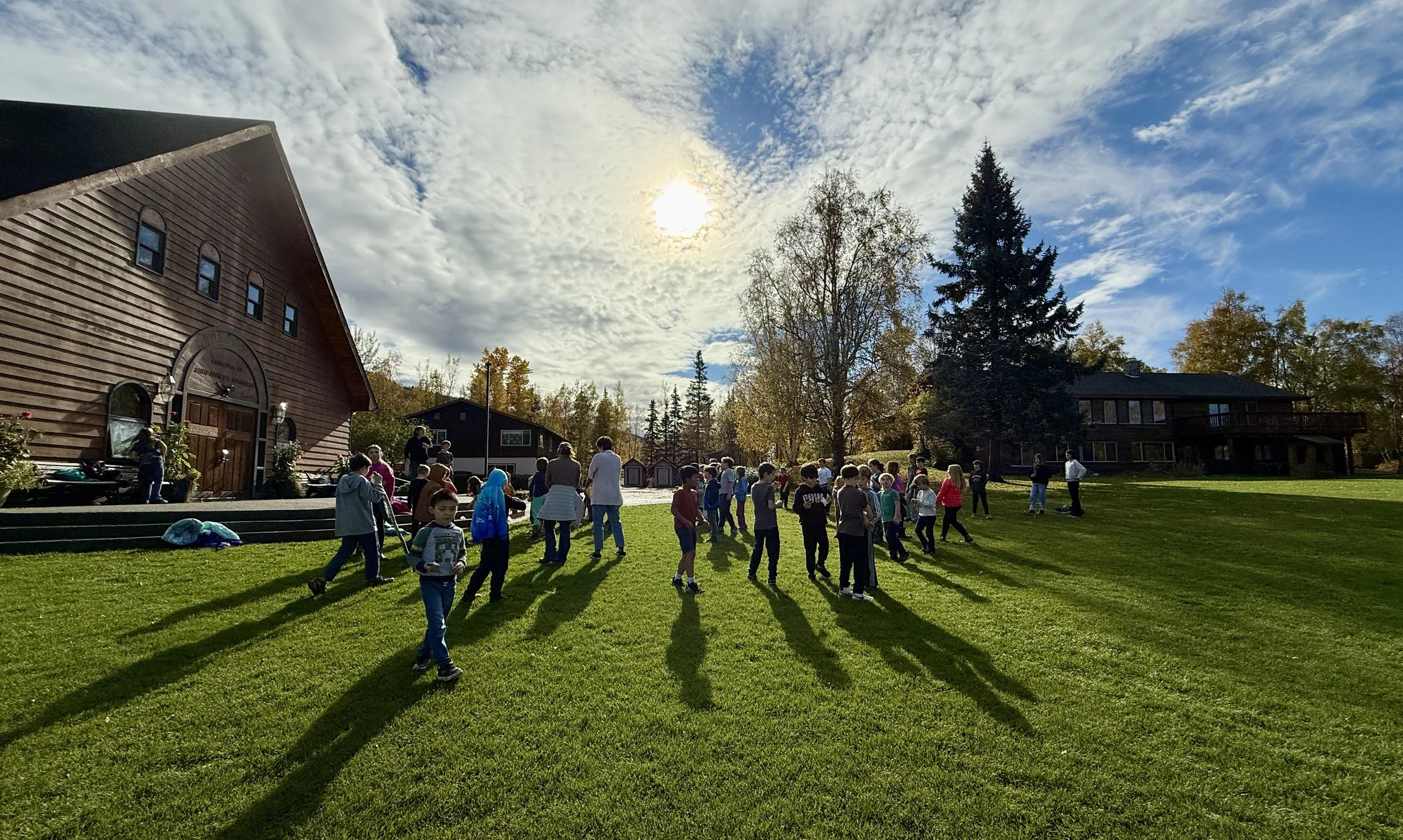 Children and adults gather on a grassy area with a large building to the left, trees in the background, and the sun partially covered by clouds in the sky.