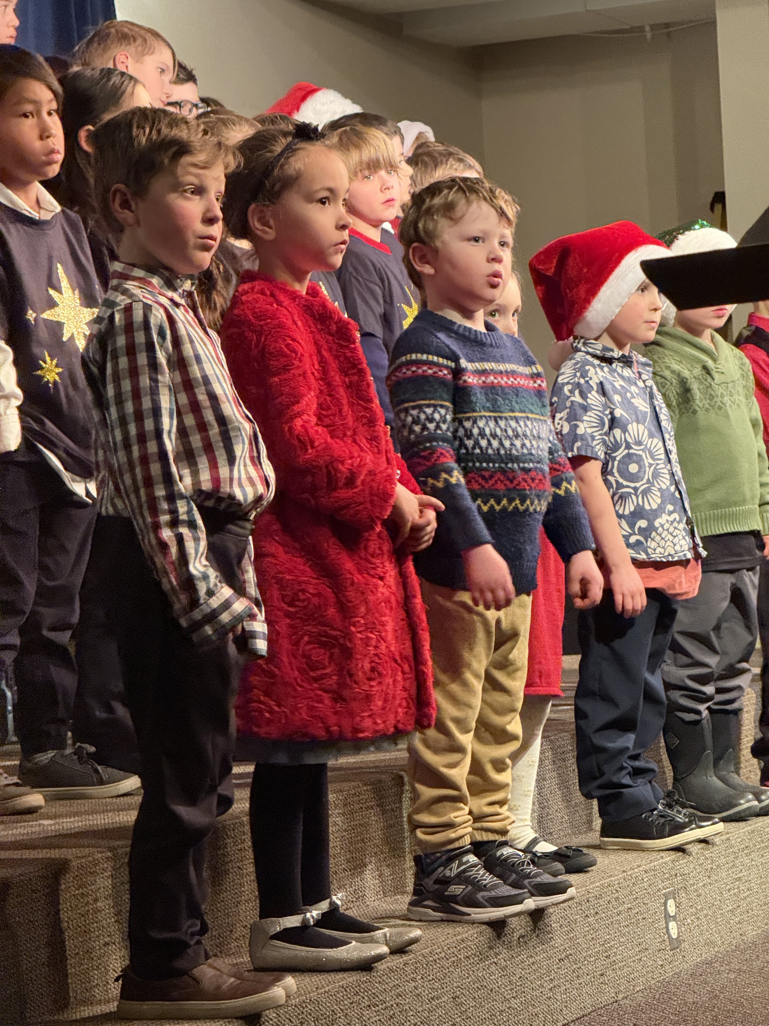 Group of children standing on a platform, singing or performing, some wearing holiday-themed clothing and accessories like Santa hats, with a focus on their feet and lower bodies.