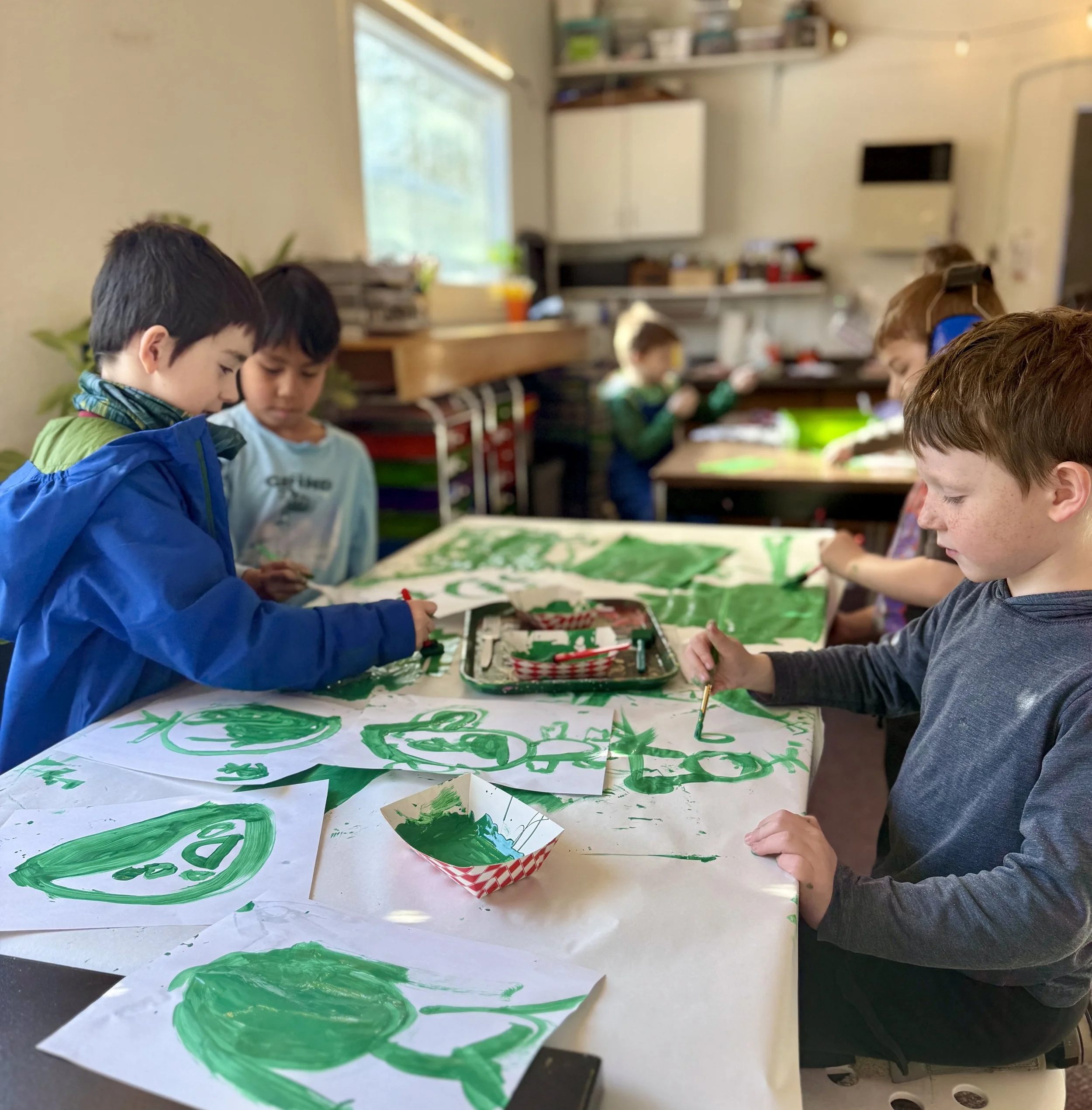 Children gathered around a table with holiday-themed crafts, green and red decorations, and artwork, in a classroom setting.