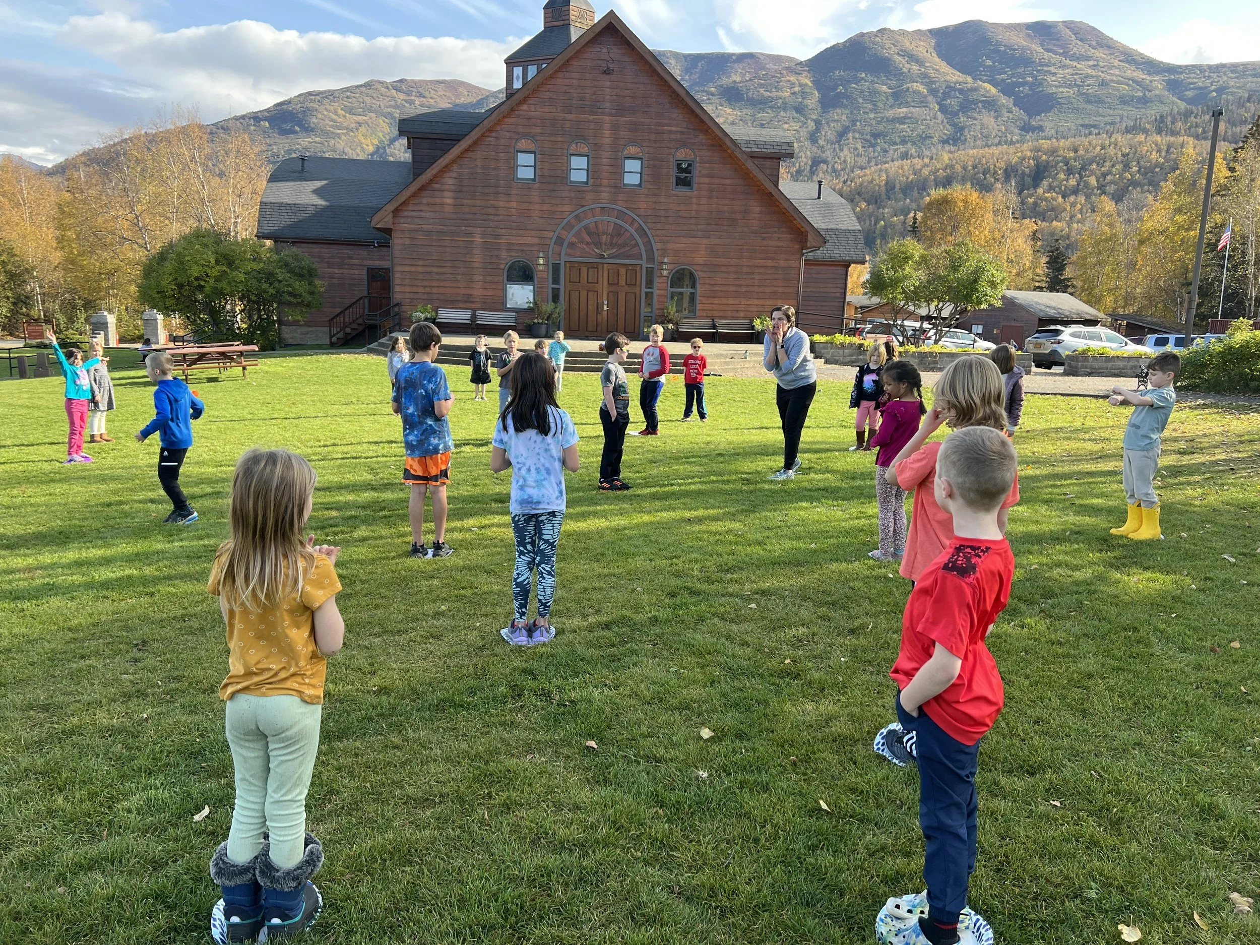 Children are playing outside on a green lawn in front of a large wooden church with mountains in the background, surrounded by trees with fall foliage.