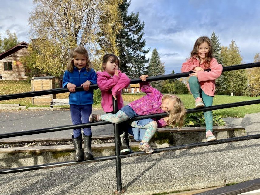 Four young girls playing and climbing on a black metal railing outdoors in a park, with trees and a house in the background on a cloudy day.