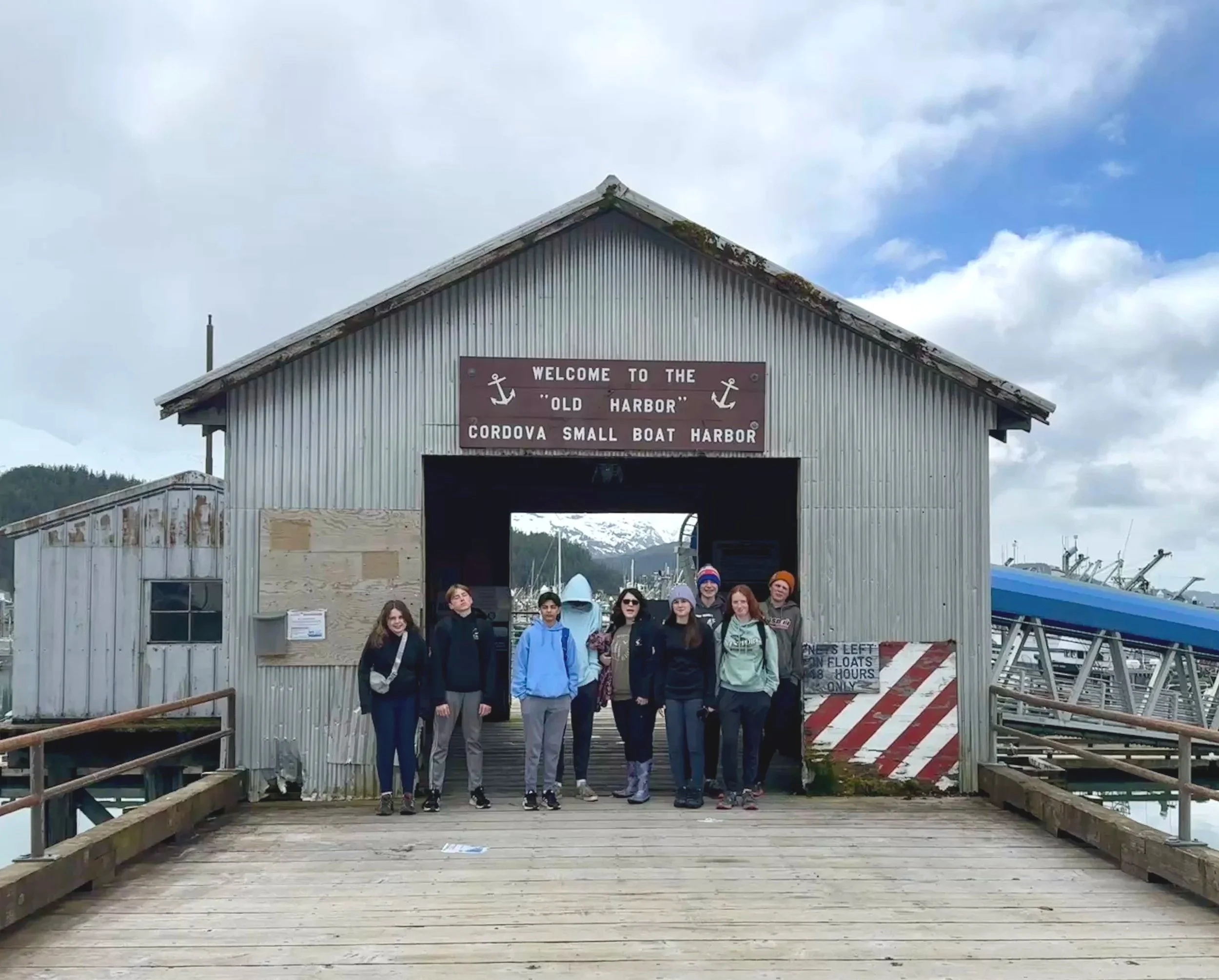 Group of people standing on a wooden dock in front of a metal building with a welcome sign at Cordova Small Boat Harbor.
