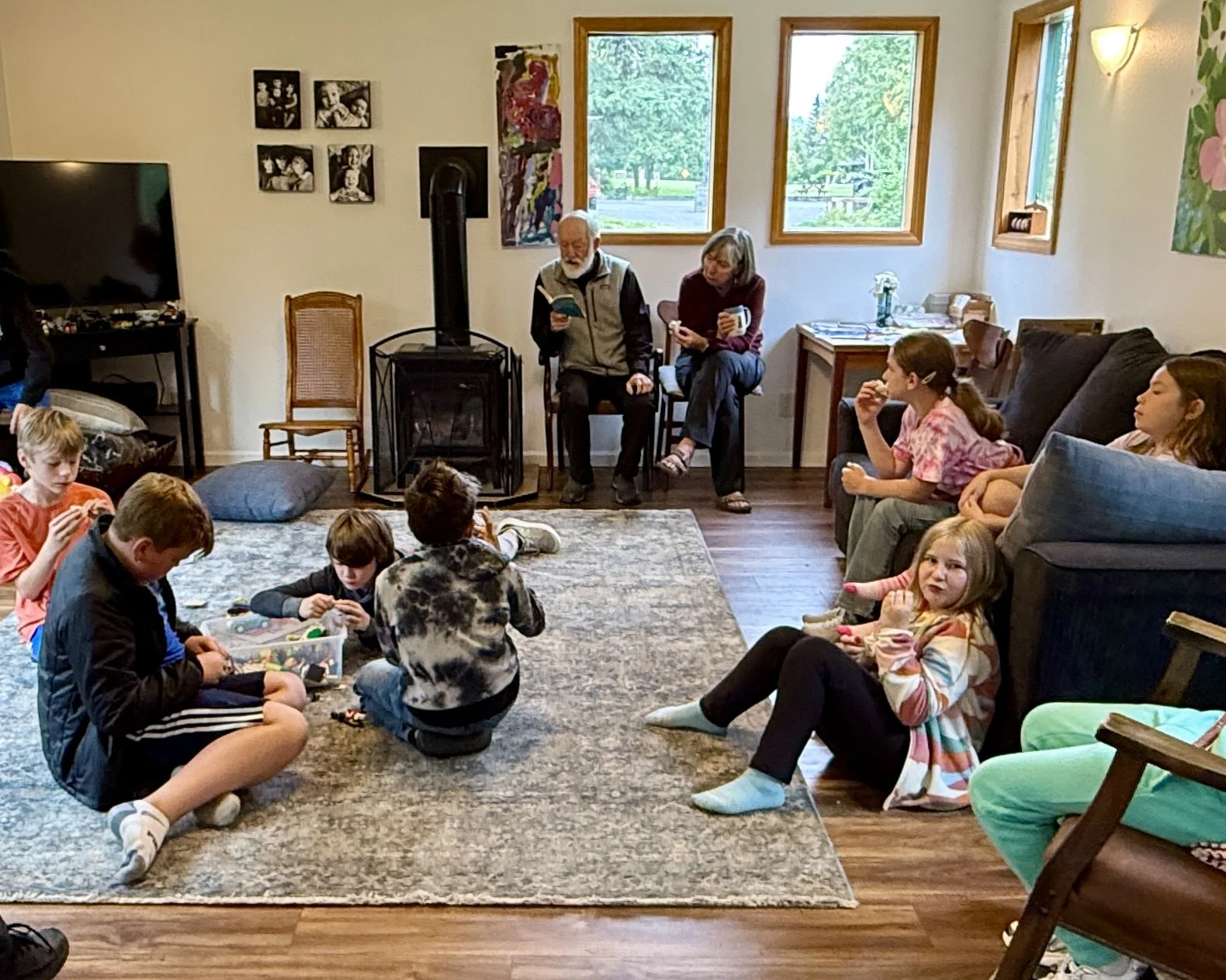 A group of children and adults gathered in a cozy living room. Children are sitting on the carpet playing with toys, while adults are seated on chairs and sofa. The room has large windows letting in natural light, decorated with artwork and family photos on the walls.