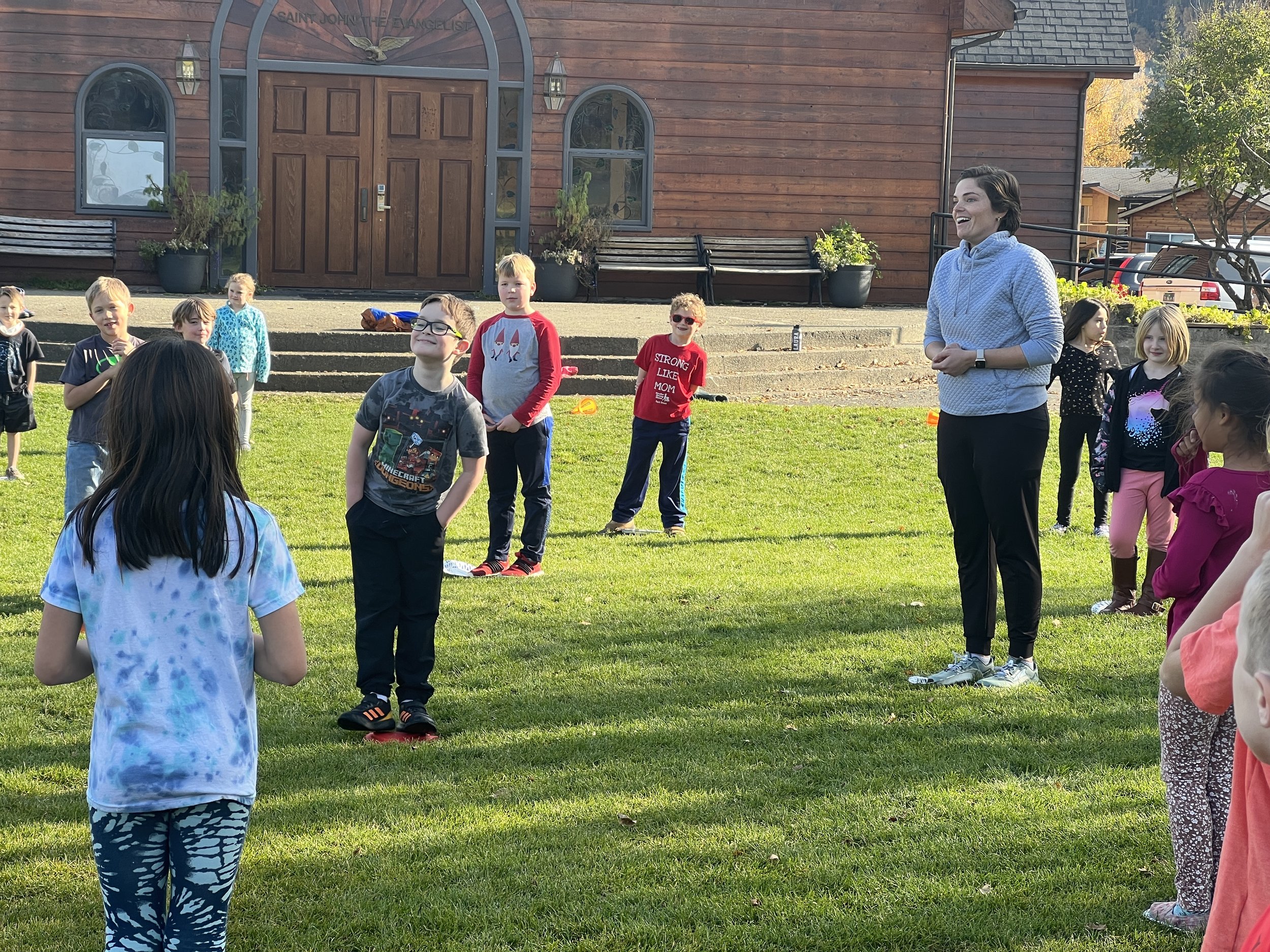 A group of children standing outdoors on a grassy area, with a woman smiling and talking to them. There is a wooden building with large doors and windows in the background.