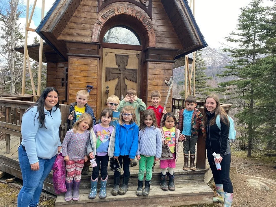 Group of children and two women standing in front of a small wooden chapel with a cross and inscriptions, amid trees in a wooded area.