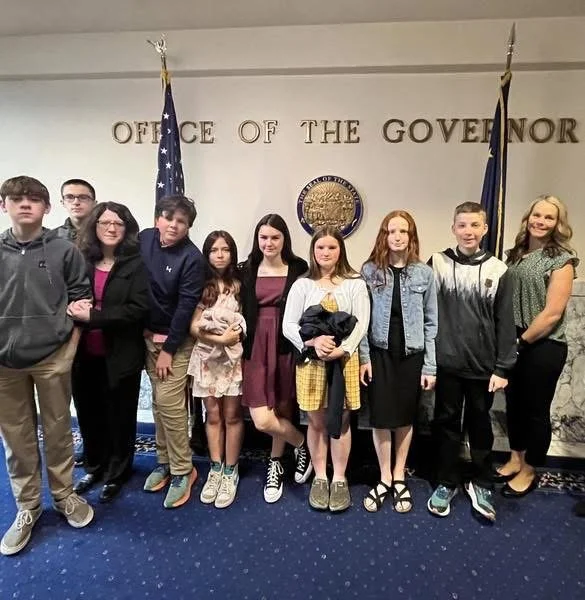 Group of children and teenagers standing in front of a wall with flags and the text 'Office of the Governor'.