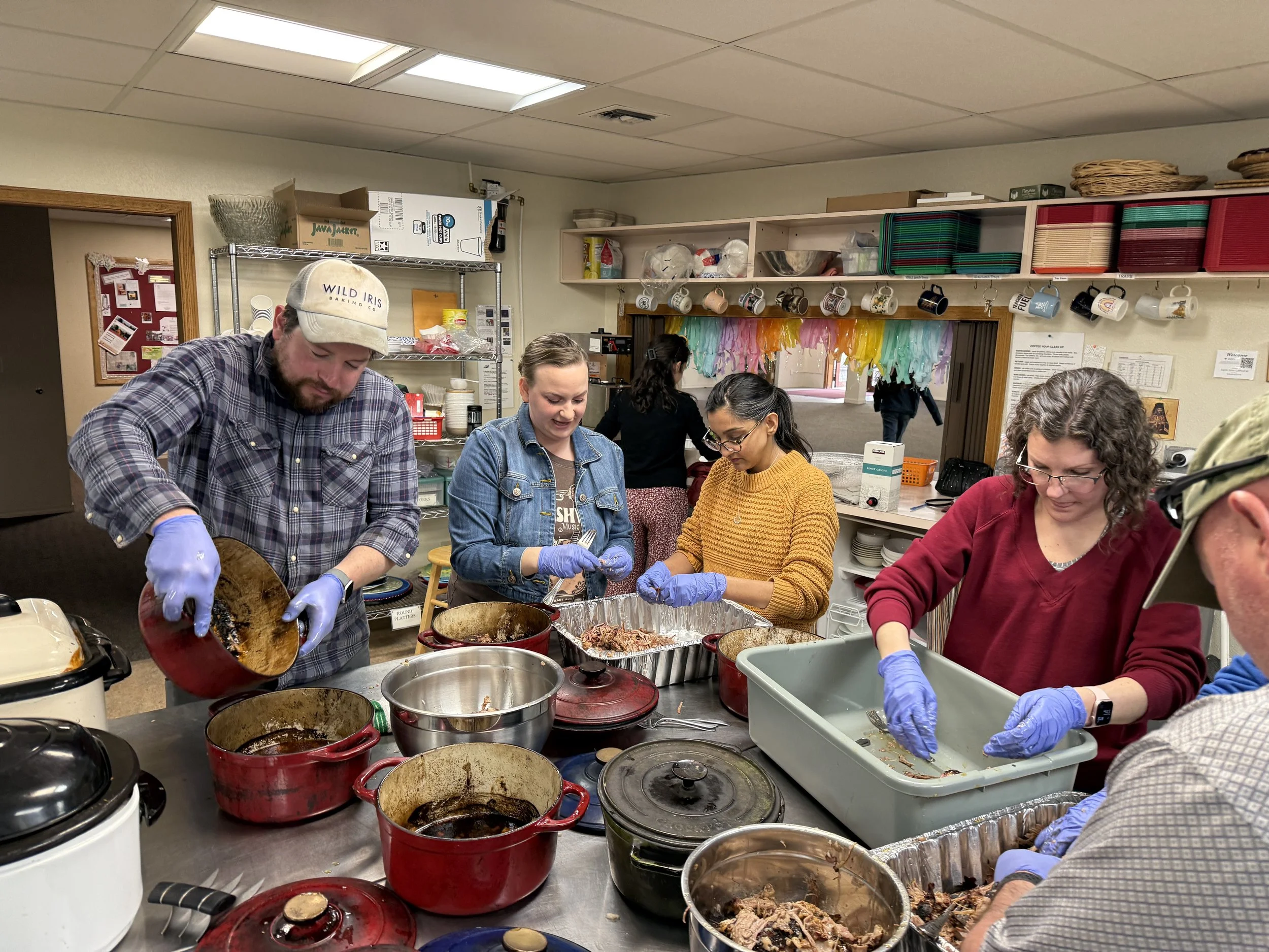 People working in a kitchen or cafeteria, preparing and serving food. Several individuals are wearing gloves and handling various dishes and containers on a counter filled with cookware, trays, and food items.