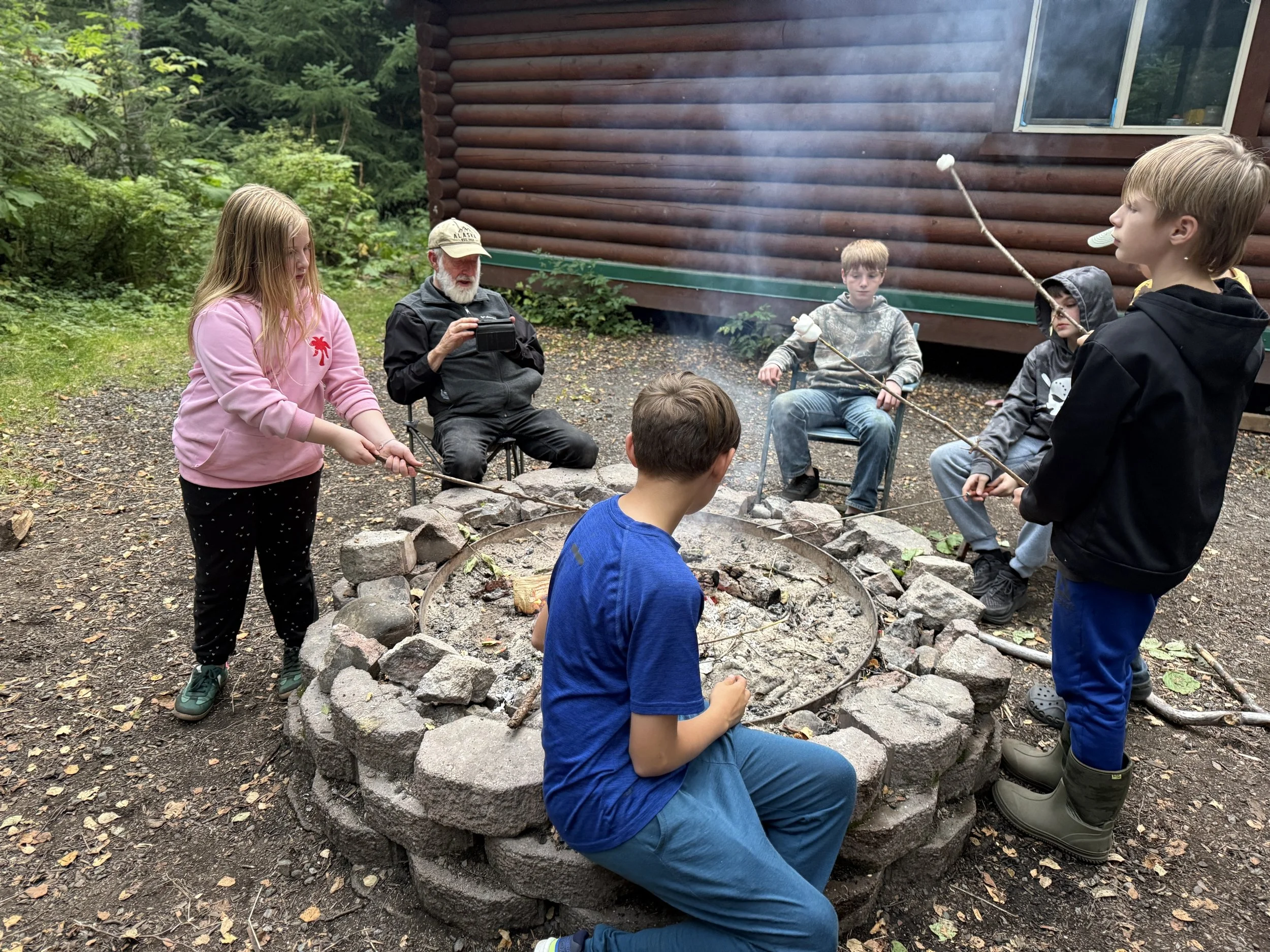 A group of children and an older man sitting outside around a fire pit, roasting marshmallows on sticks near a log cabin in the woods.