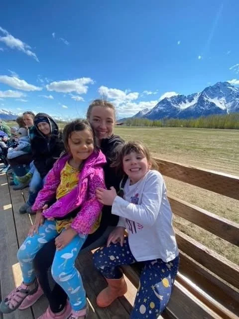 Group of children and a young woman sitting on a wooden bench outdoors with mountains and blue sky in the background