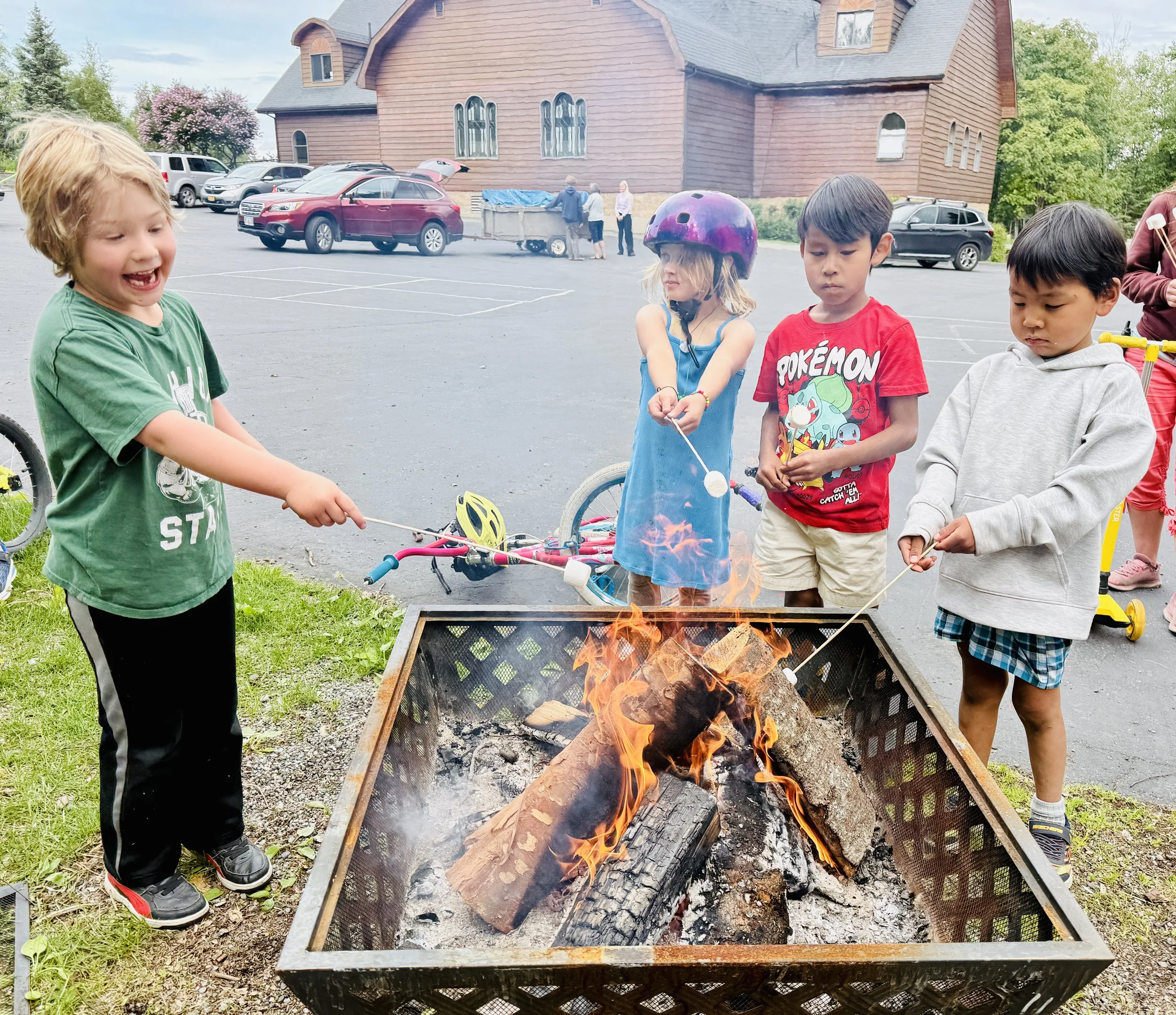 Children gathered around a fire pit outdoors, roasting marshmallows. A large brick building and parked cars are in the background.