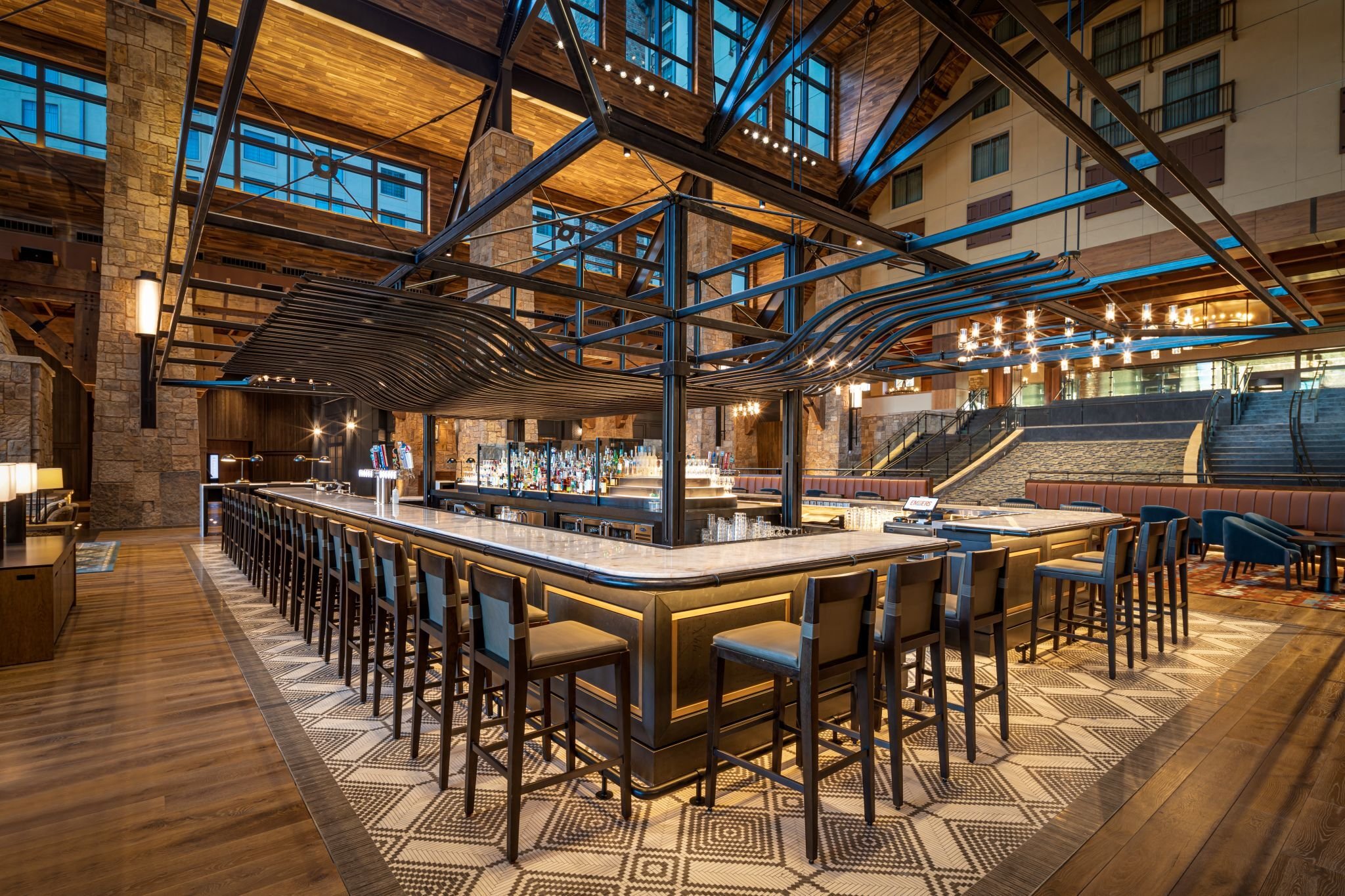 Interior of a modern hotel lobby or bar area with a large curved bar counter, high chairs, a chandelier, and a multi-level structure with windows and stone and wood accents.