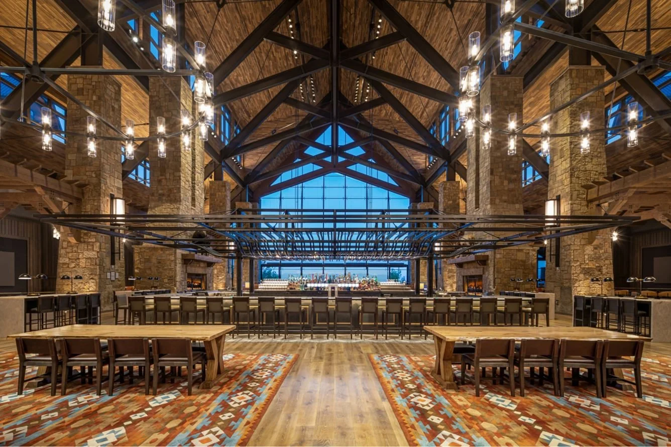 Interior of a large, rustic-style bar or restaurant with high vaulted wooden ceiling, stone columns, and modern chandeliers. A bar with barstools is at the back, and seating with wooden tables and chairs is at the front. The space has large windows and warm lighting.