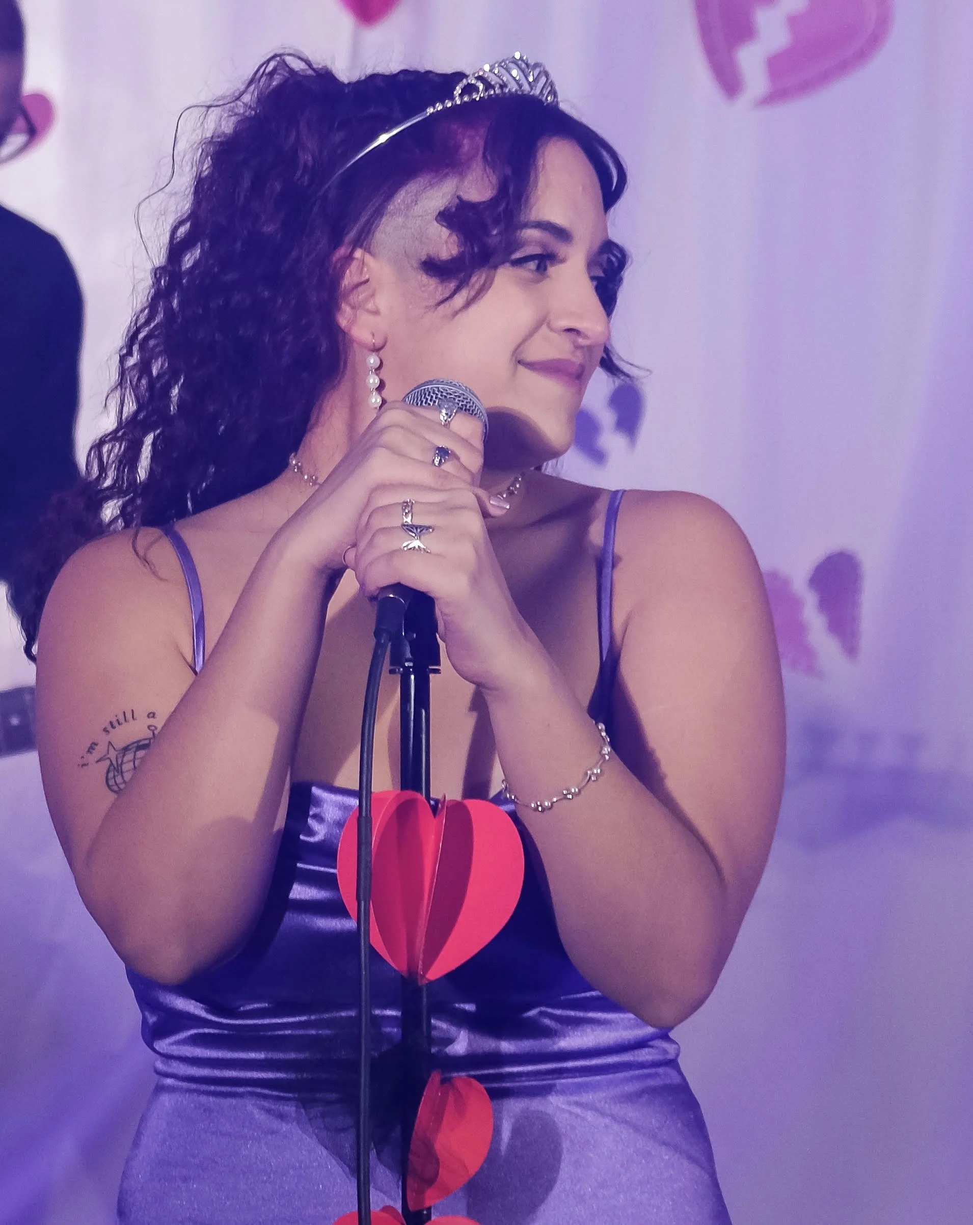 A woman with curly dark hair wearing a small crown, pearl earrings, a necklace, and a dress holding a microphone, smiling, with a purple heart in the background.