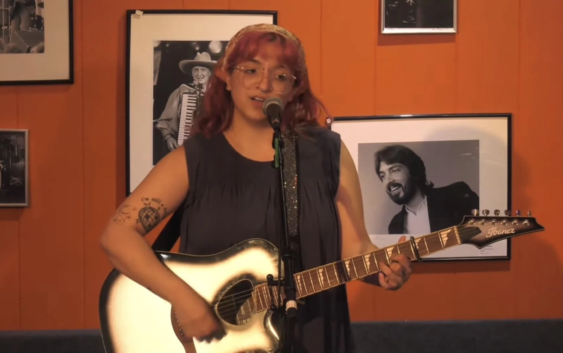 A woman with red hair and glasses playing an acoustic guitar and singing into a microphone in front of a red wall decorated with black and white framed photographs.