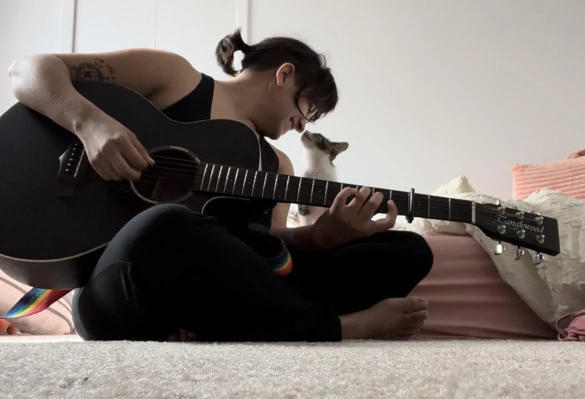 A person sitting cross-legged on a bed playing an acoustic guitar, with a cat looking at them.