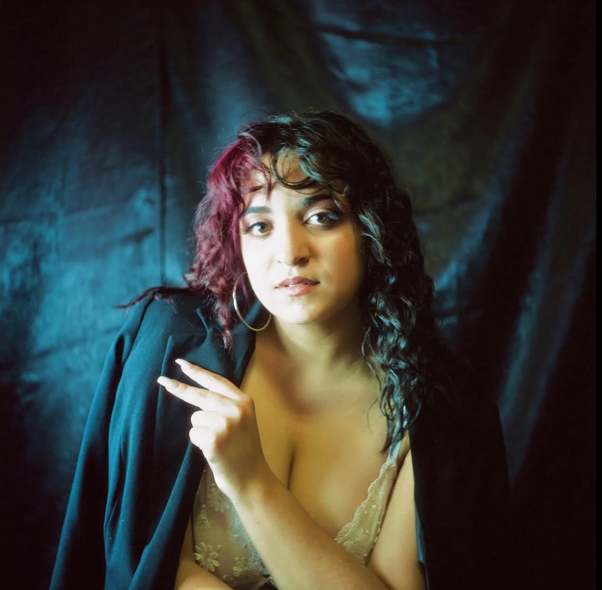 A woman with curly, dark hair and hoop earrings looking at the camera. She has light skin and is wearing a lace top with a black jacket draped over her shoulders. She is posed with her right hand near her collarbone and pointing with her index finger.