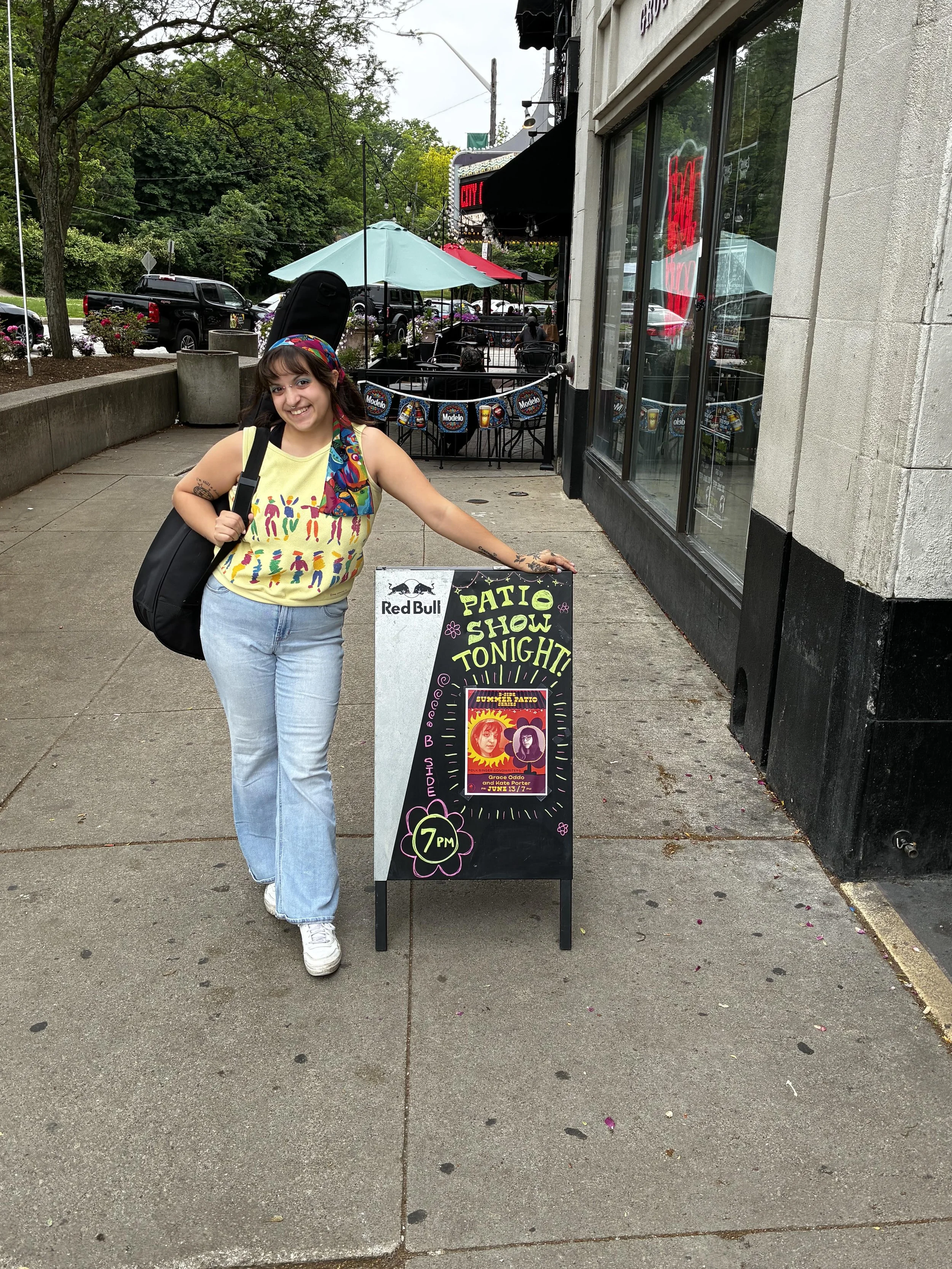 A smiling woman with a tattoo on her arm, wearing a colorful headband and a bright yellow tank top with colorful figures, stands on a sidewalk outside a restaurant. She is carrying a black bag over her shoulder. In front of her is a chalkboard sign advertising a patio show night at 7 PM with a poster for a musical event featuring artists Grace Odo and Kate Porter. The background shows outdoor seating with umbrellas, string lights, parked cars, trees, and a building with large windows.
