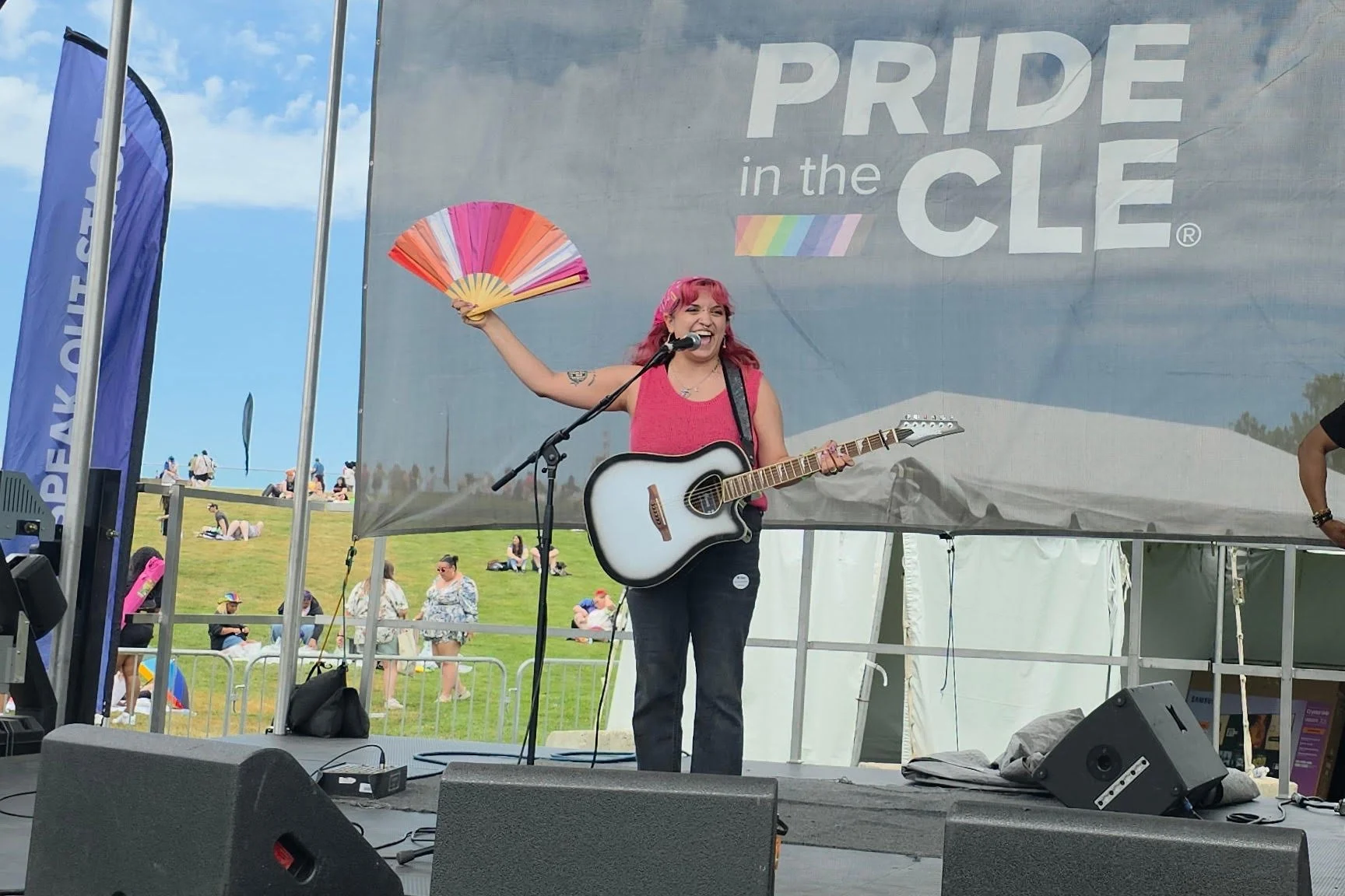 A woman with pink hair singing and playing the guitar on stage at the Pride in the Clee event, holding a colorful fan in one hand and smiling. The stage has a large banner that reads 'PRIDE in the CLEE' with a rainbow flag design.