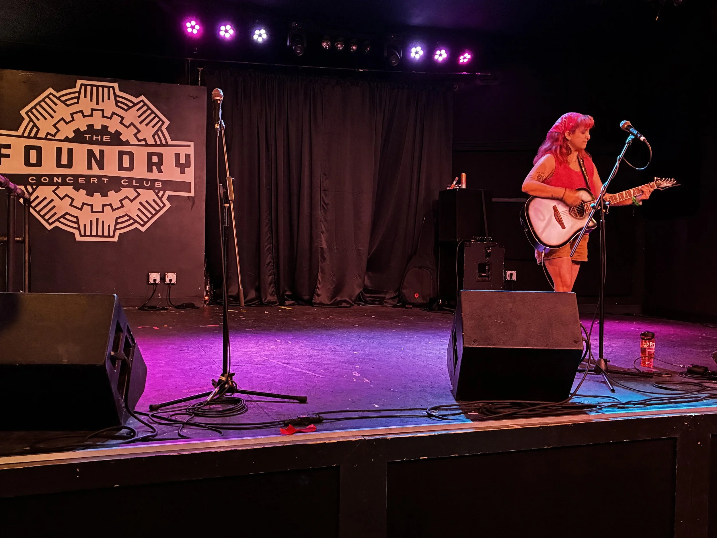 Woman with red hair playing an acoustic guitar on stage at The Foundry concert club, with microphone stands and stage lighting overhead.